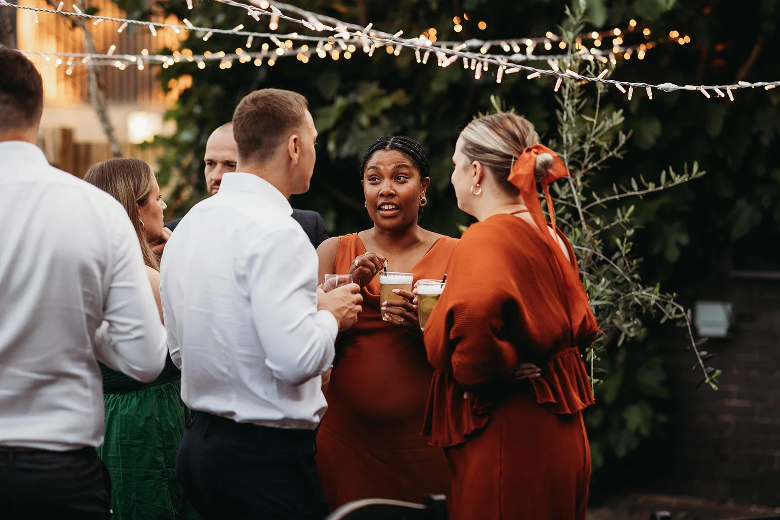 Group of people talking and holding drinks at an outdoor evening party, with string lights overhead and greenery in the background.