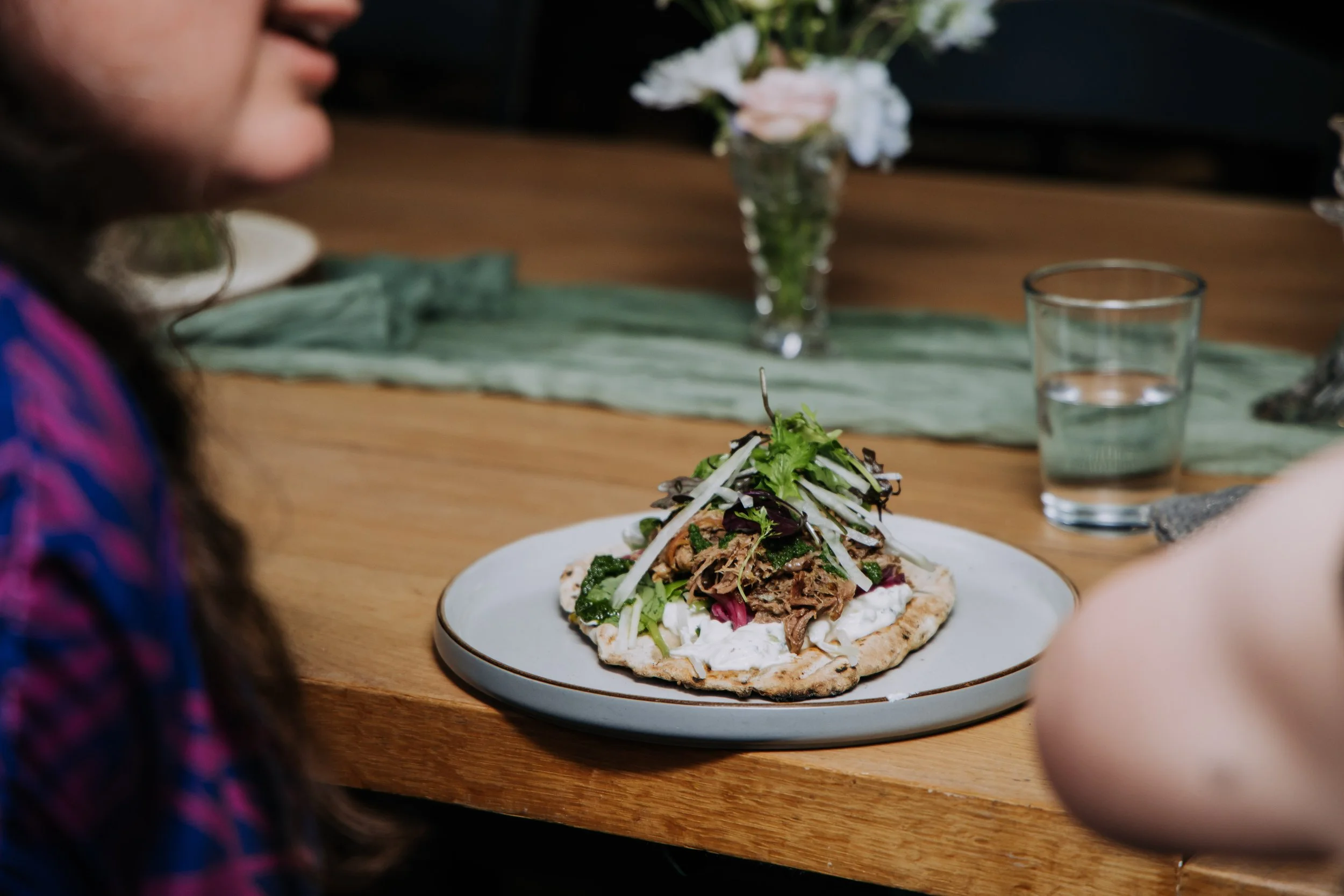 A flatbread topped with shredded meat, greens, and vegetables on a white plate, on a wooden table with a glass of water and a vase with flowers in the background.