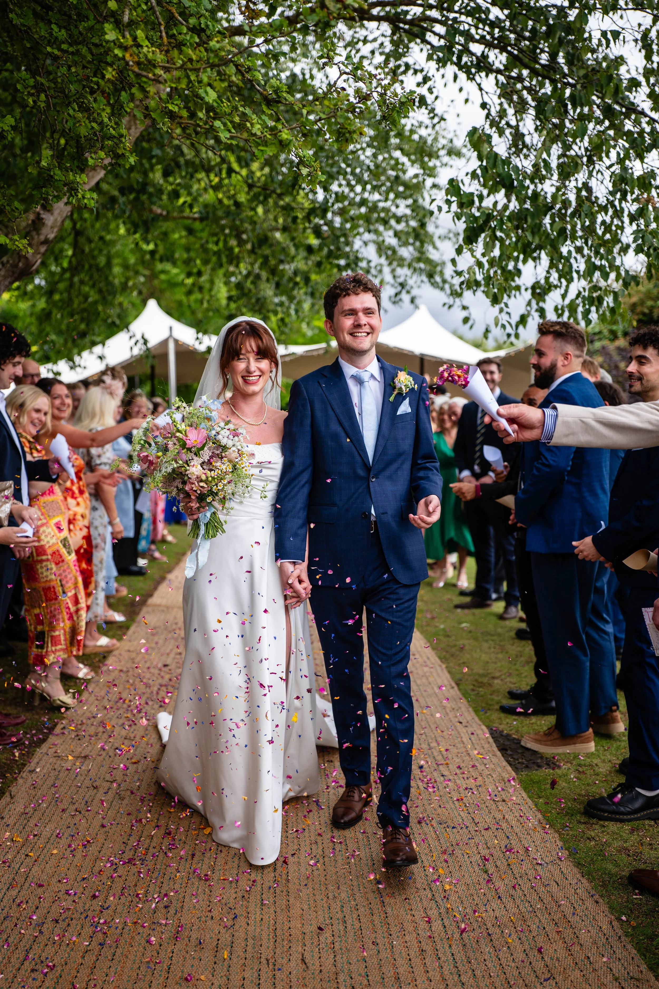 A newlywed couple walking down an outdoor aisle, holding hands, surrounded by friends and family throwing confetti. The bride is in a white wedding dress holding a bouquet, and the groom is in a blue suit with a boutonniere. Green trees and white ten