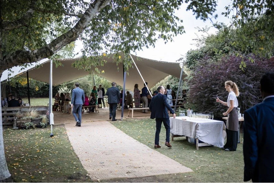 Outdoor wedding reception with a large tent, guests mingling, and a table with champagne glasses and a server.