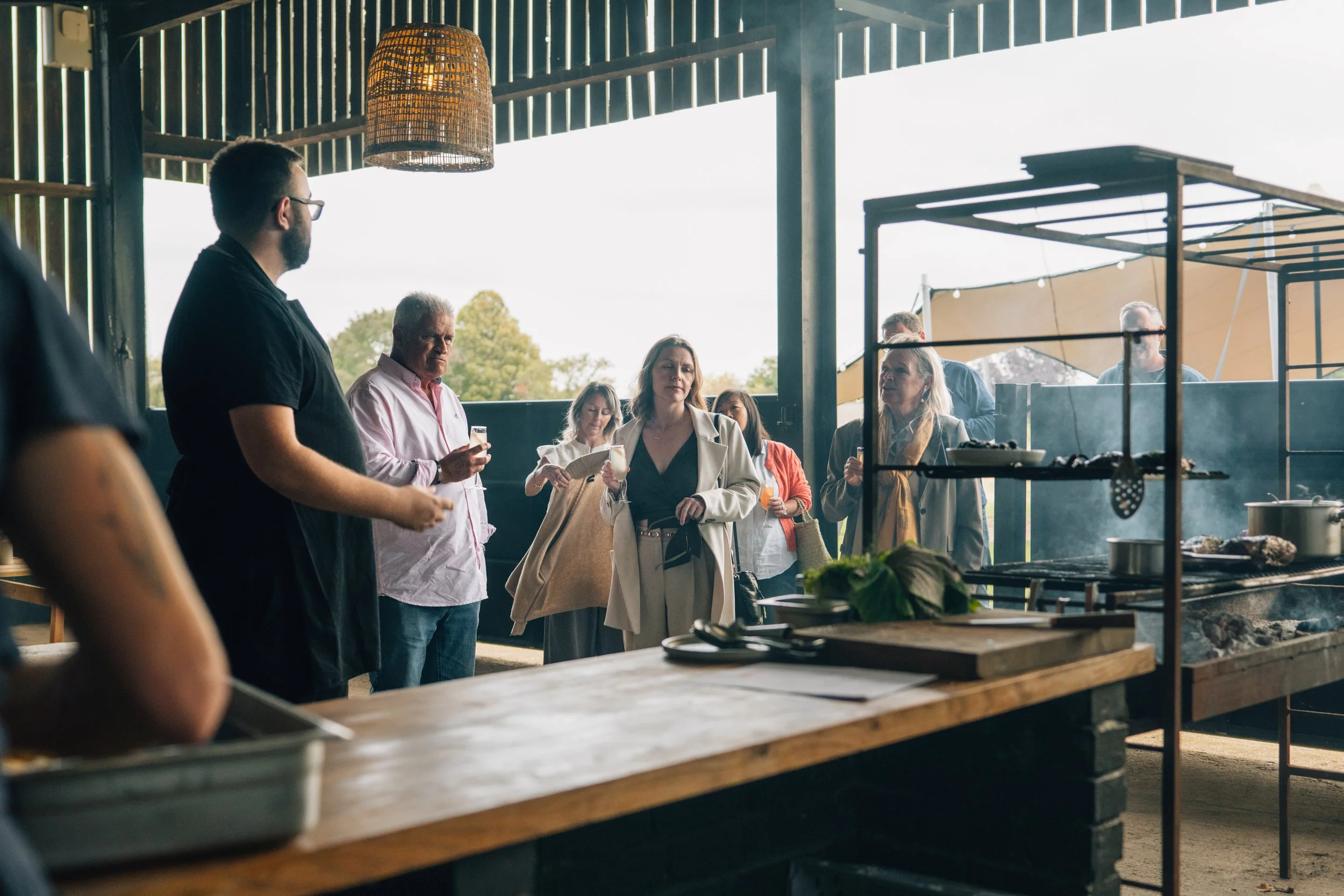 People gathered at an outdoor food stall or restaurant with a grill, some are eating and using their phones, in a semi-open structure with wooden and metal elements, during daytime.