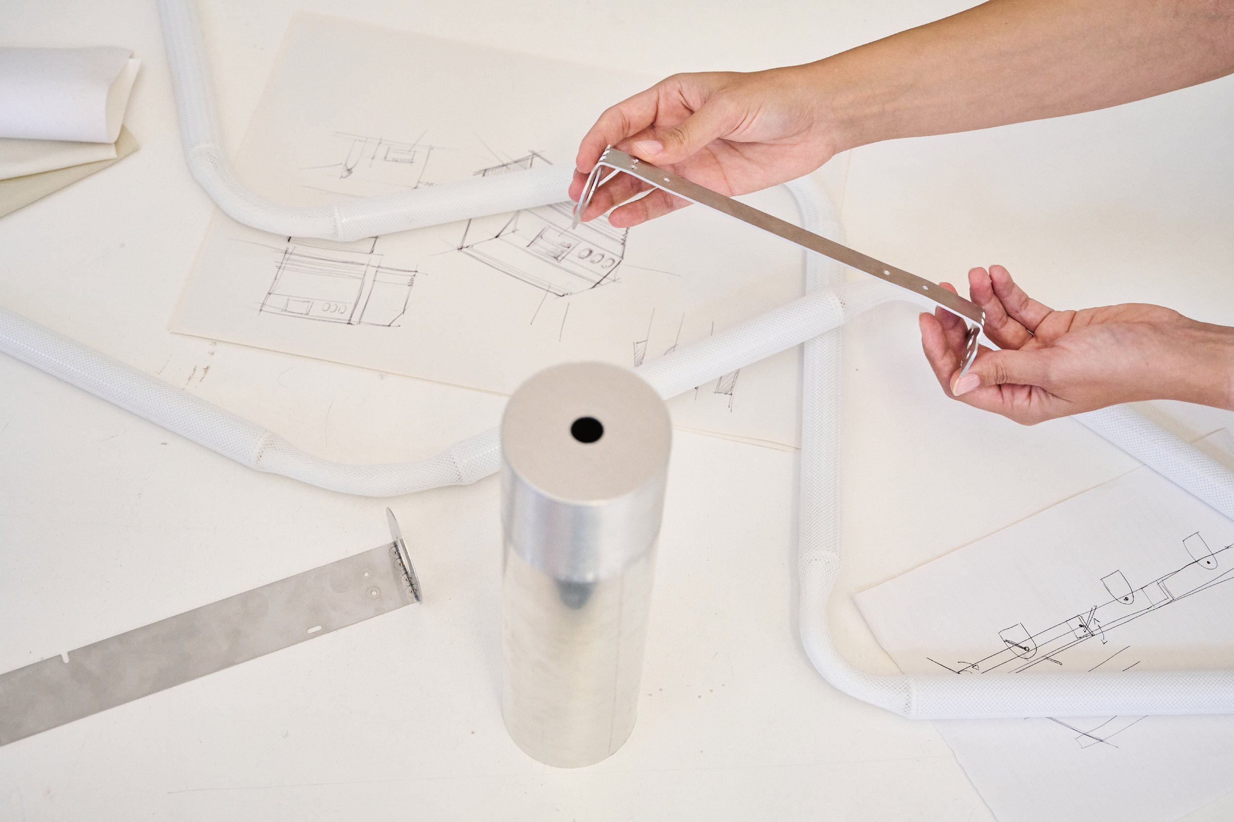 Hands holding a metal bracket over a table with sketches and metal components, including pipes and a cylindrical structure.