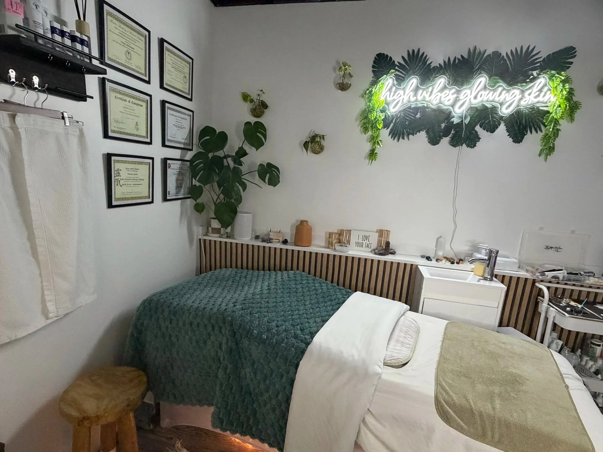 A cozy bedroom with framed certificates on the wall, a bed with white and green blankets, a white cabinet with plants and decorative items, and a neon sign on the wall that says 'good vibes only,' with a small rainbow light reflection.