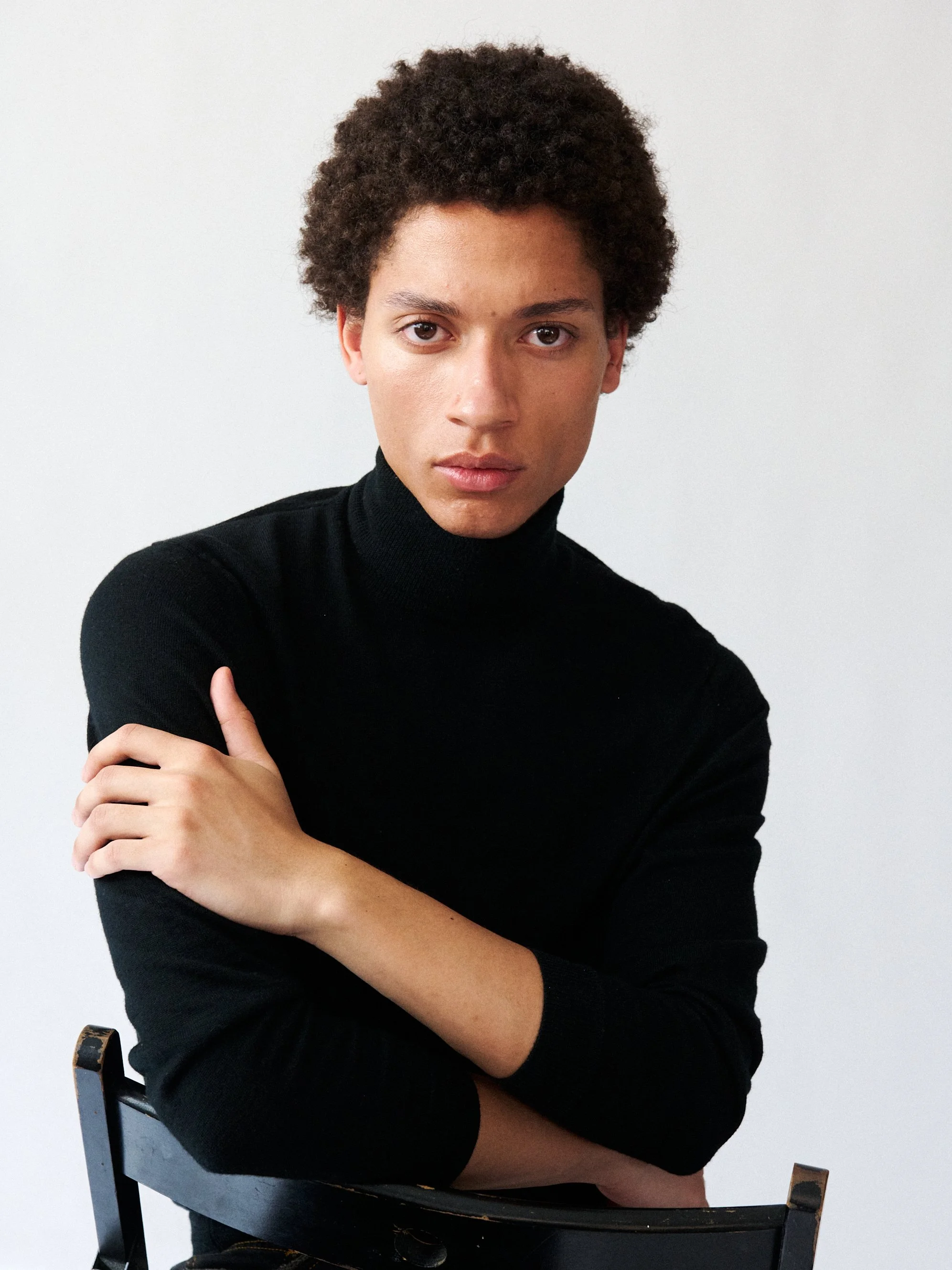 Male model with an afro wearing a black turtleneck sitting on a black chair and crossing his arms, looking directly at the camera with a neutral expression against a plain light background.