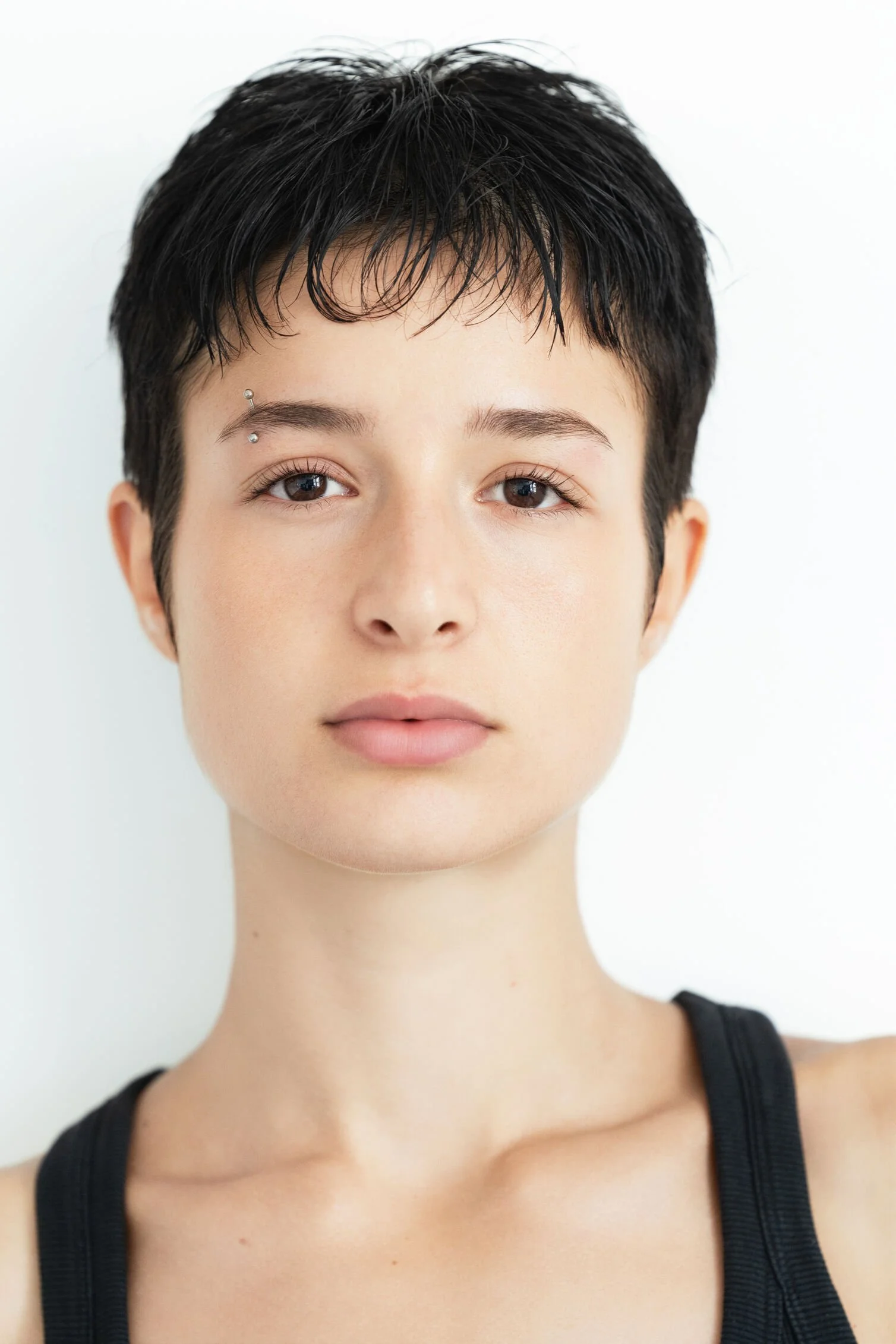 A close-up portrait of a young female model with short, dark, wet hair, light skin, brown eyes, and a facial piercing above her left eyebrow. She is wearing a black tank top and standing against a plain white background.