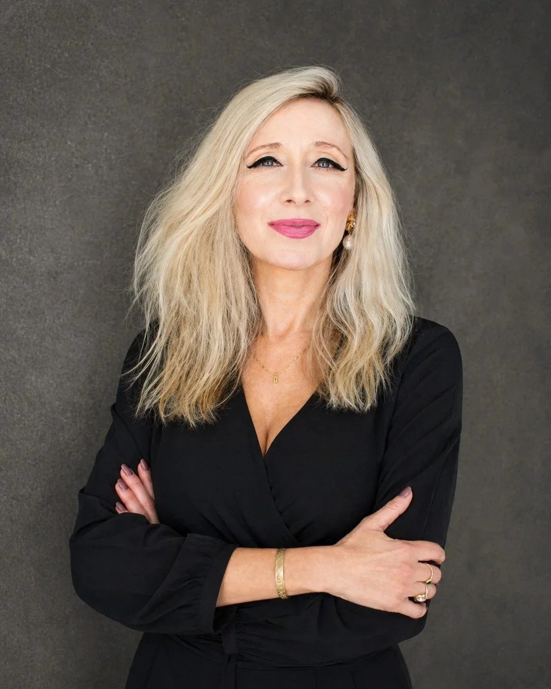 A woman with long blonde hair, wearing a black dress, gold jewelry, and pink lipstick, standing against a gray background.