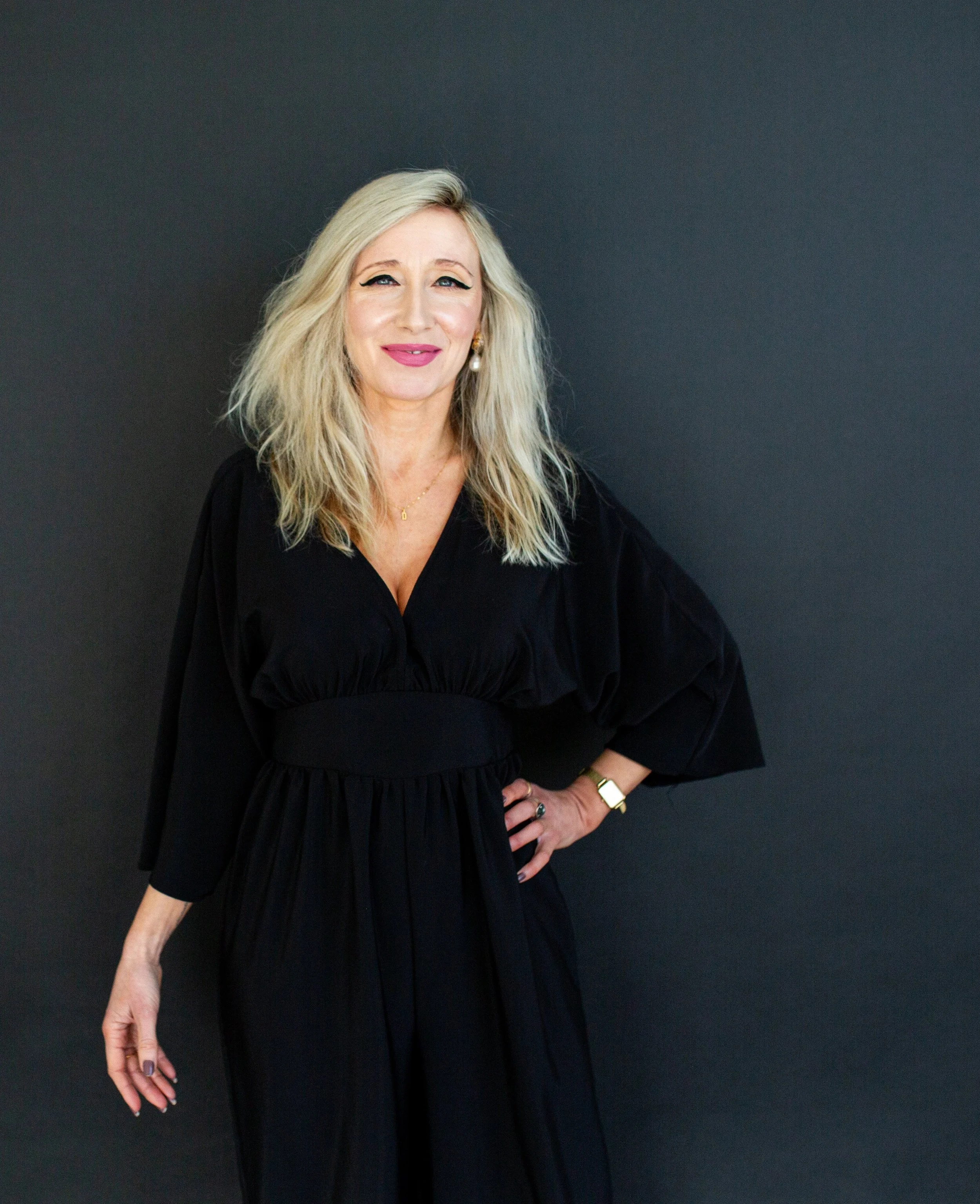 A woman with long blonde hair and light makeup standing against a dark background, wearing a black dress, gold jewelry, and a gold watch.