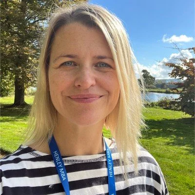 A woman with blonde hair and blue eyes smiling outdoors. She is wearing a black-and-white striped shirt and a blue lanyard, with trees, a grassy area, and a lake in the background under a partly cloudy sky.