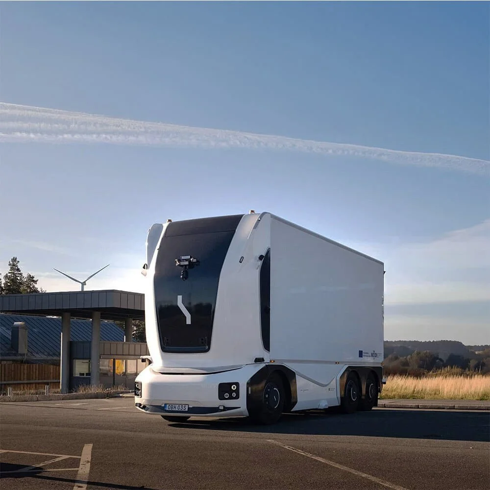A futuristic, self-driving delivery truck parked in a lot with a building, wind turbine, and grassy field in the background under a clear sky.
