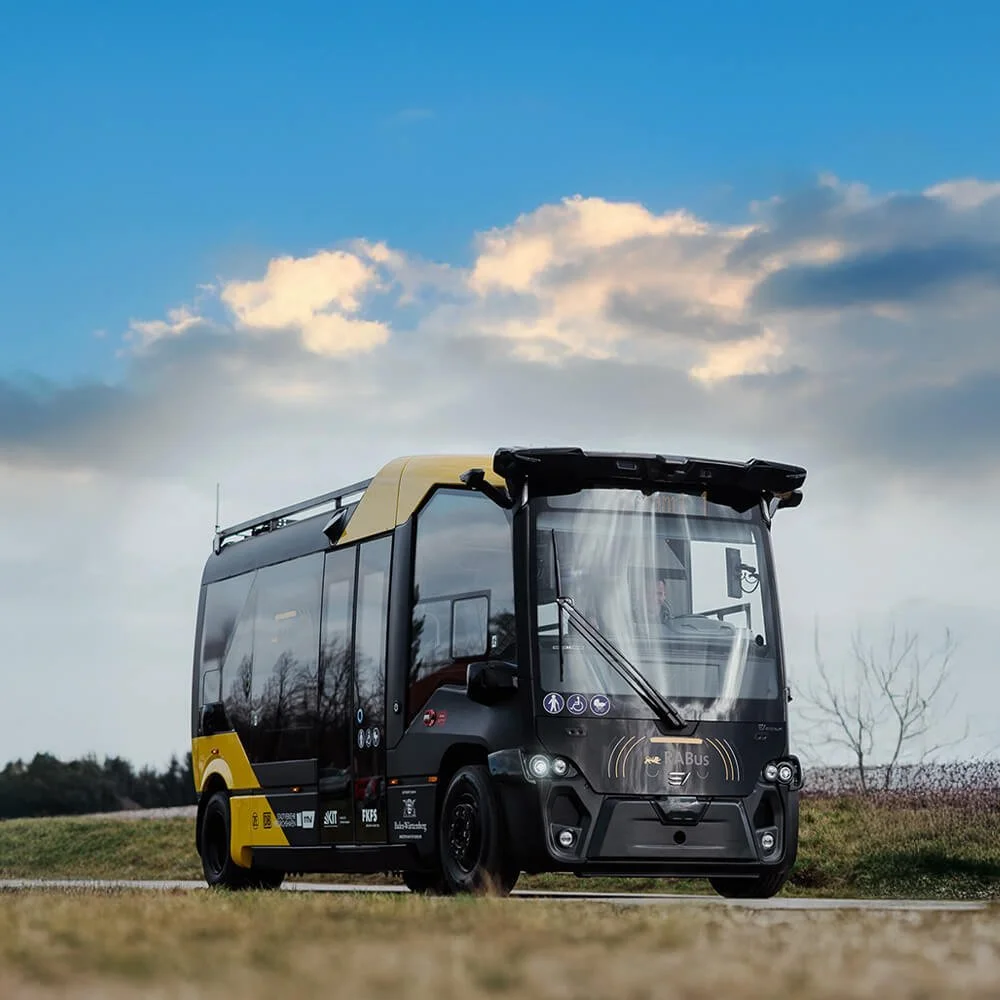 Autonomous electric shuttle bus driving on a road with clouds and a blue sky in the background.