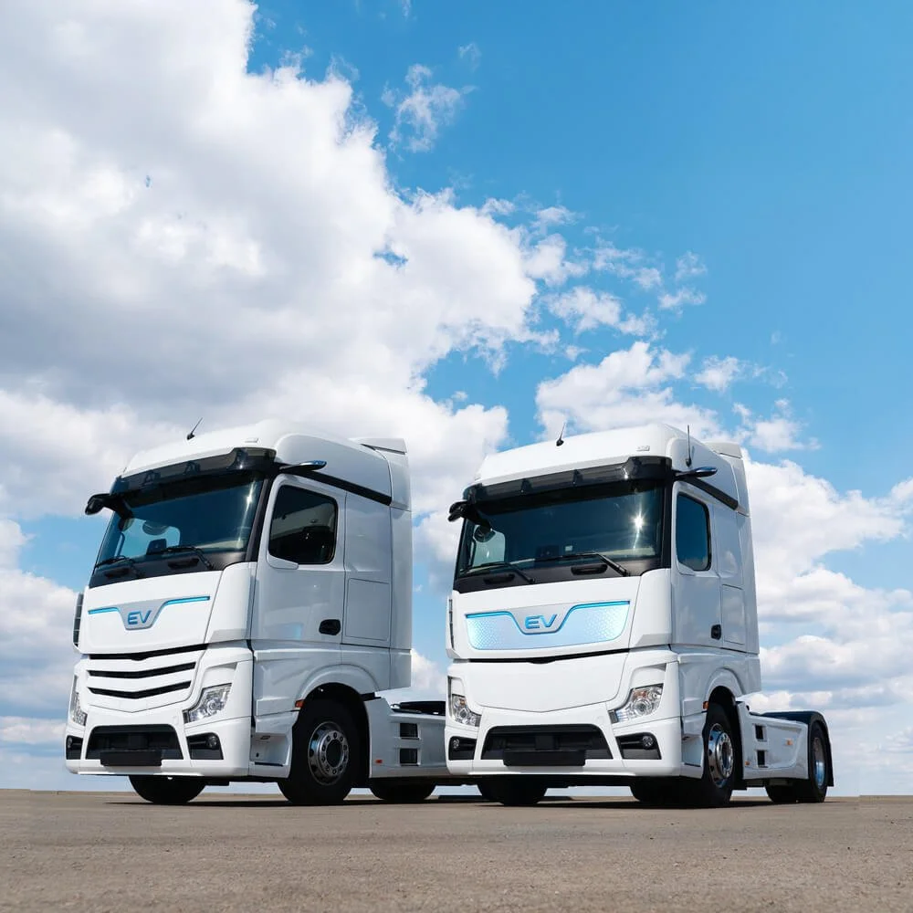 Two white electric semi-trucks parked on a flat surface under a partly cloudy sky.