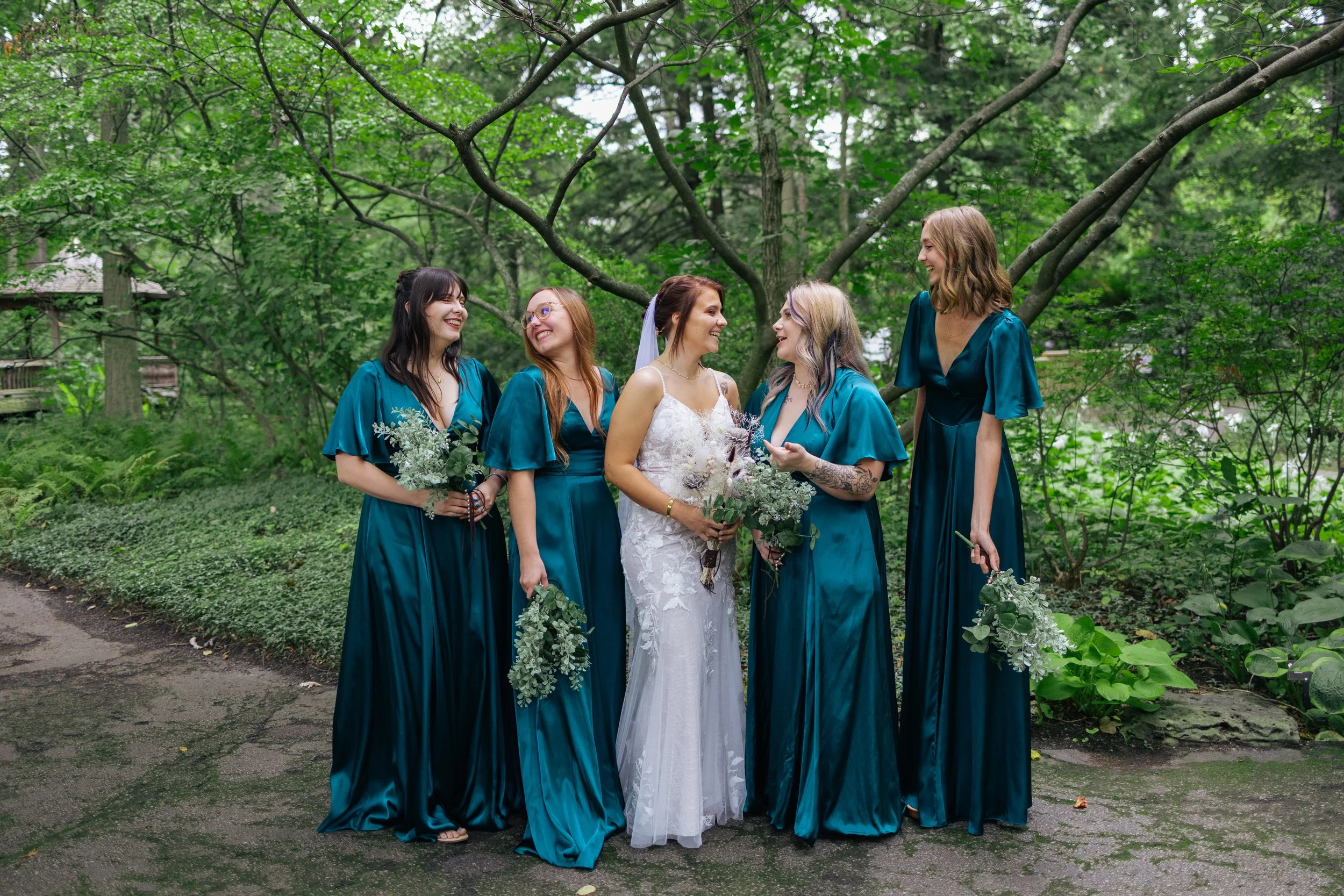A bride and her five bridesmaids in teal dresses standing outdoors in a green garden, smiling and holding bouquets, with trees and foliage in the background.