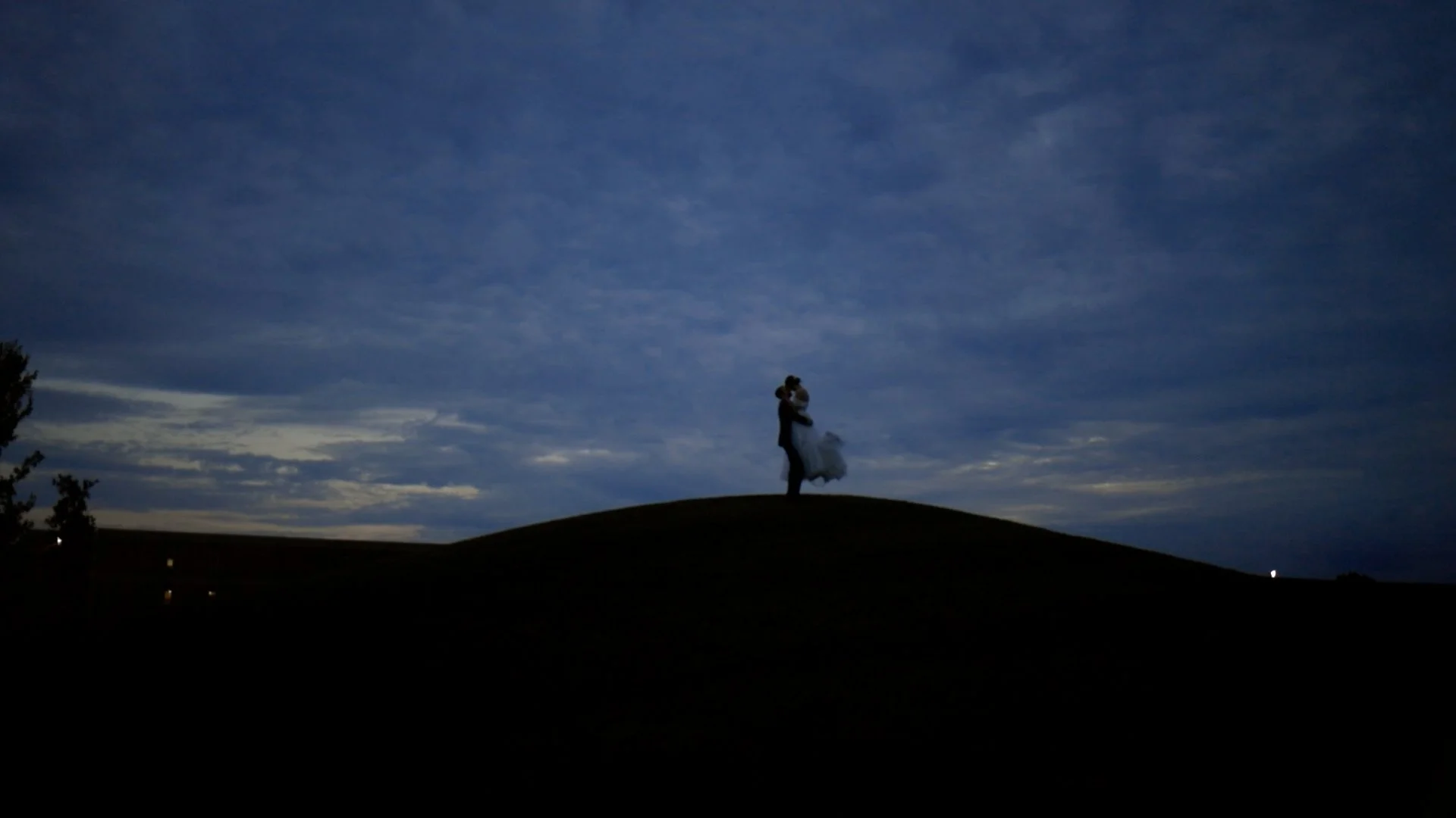 Silhouette of a couple, with the man lifting the woman in a wedding dress, standing on a hill at dusk with a cloudy sky in the background.