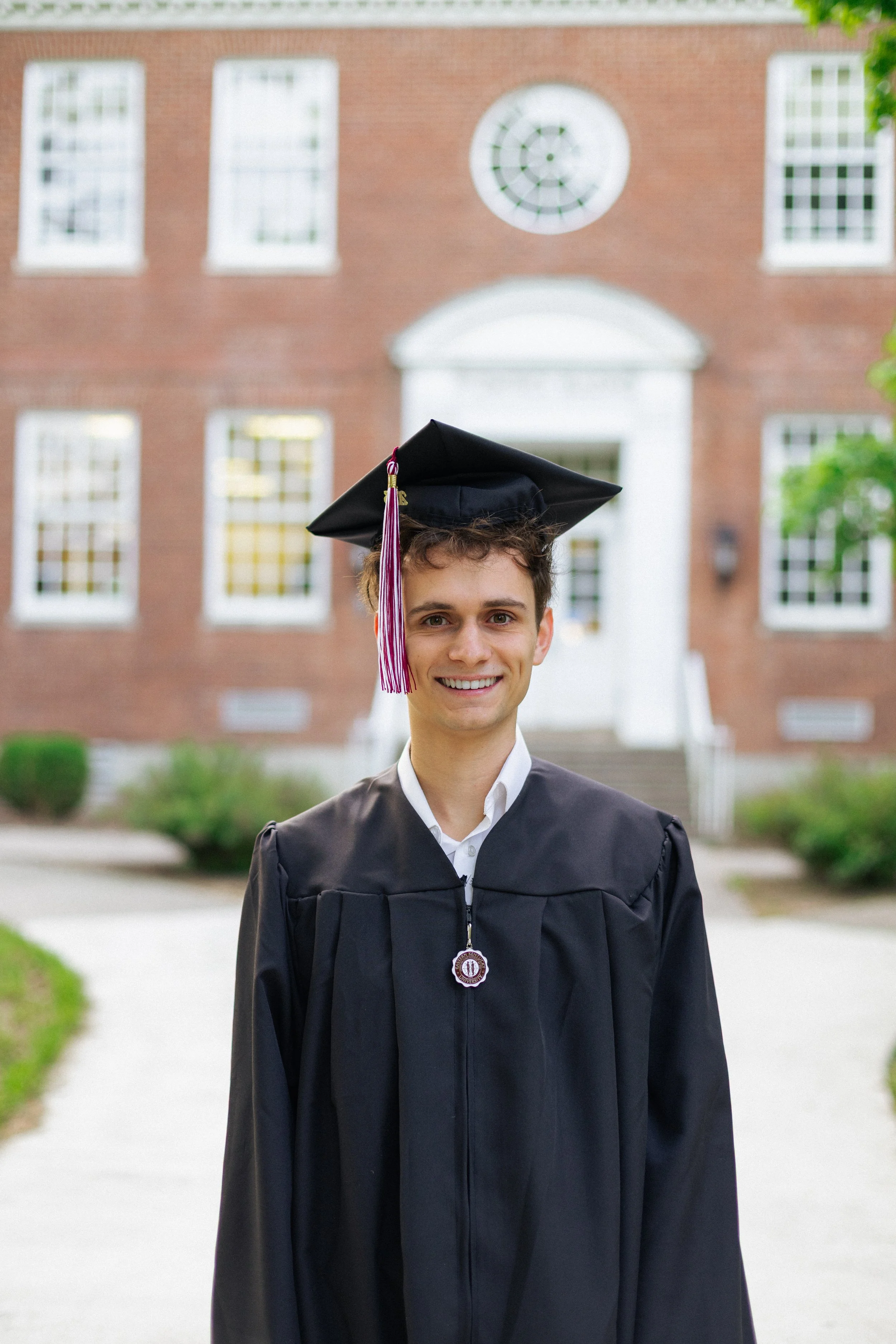 Young man in graduation cap and gown standing on a college campus walkway, smiling in front of a brick university building.