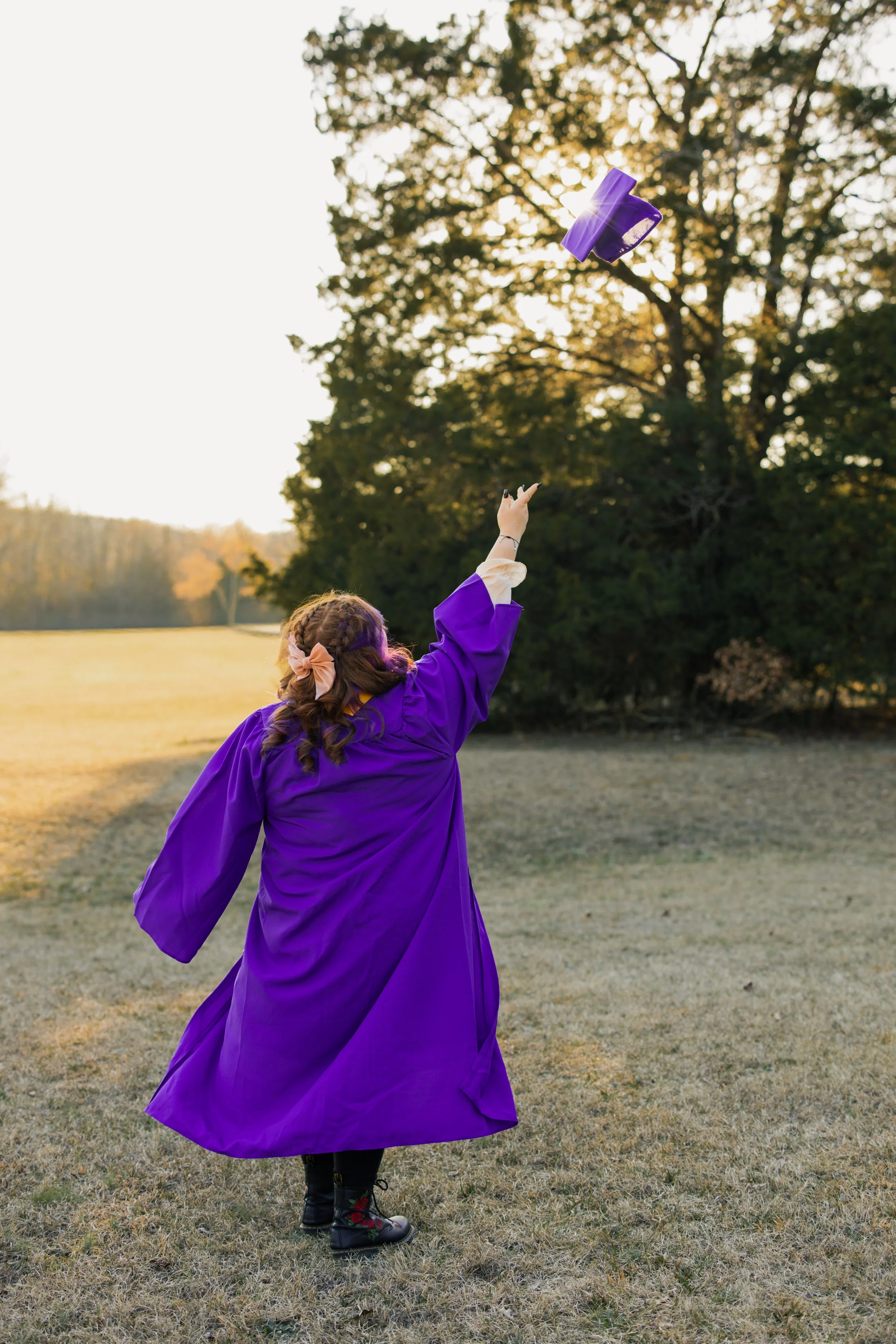 A person wearing a purple graduation gown and black boots standing in an open grassy field, throwing a purple graduation cap into the air during sunset.