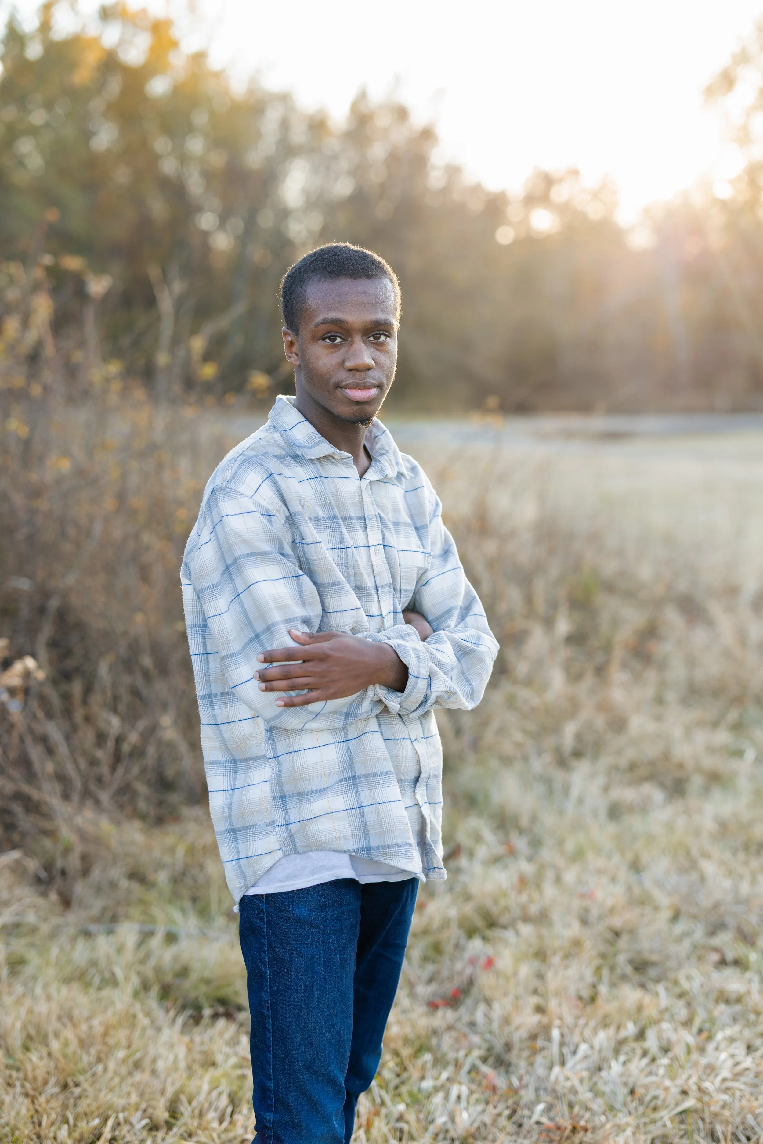 A young man standing outdoors in a field during sunset, wearing a plaid shirt and jeans, with arms crossed and looking at the camera.