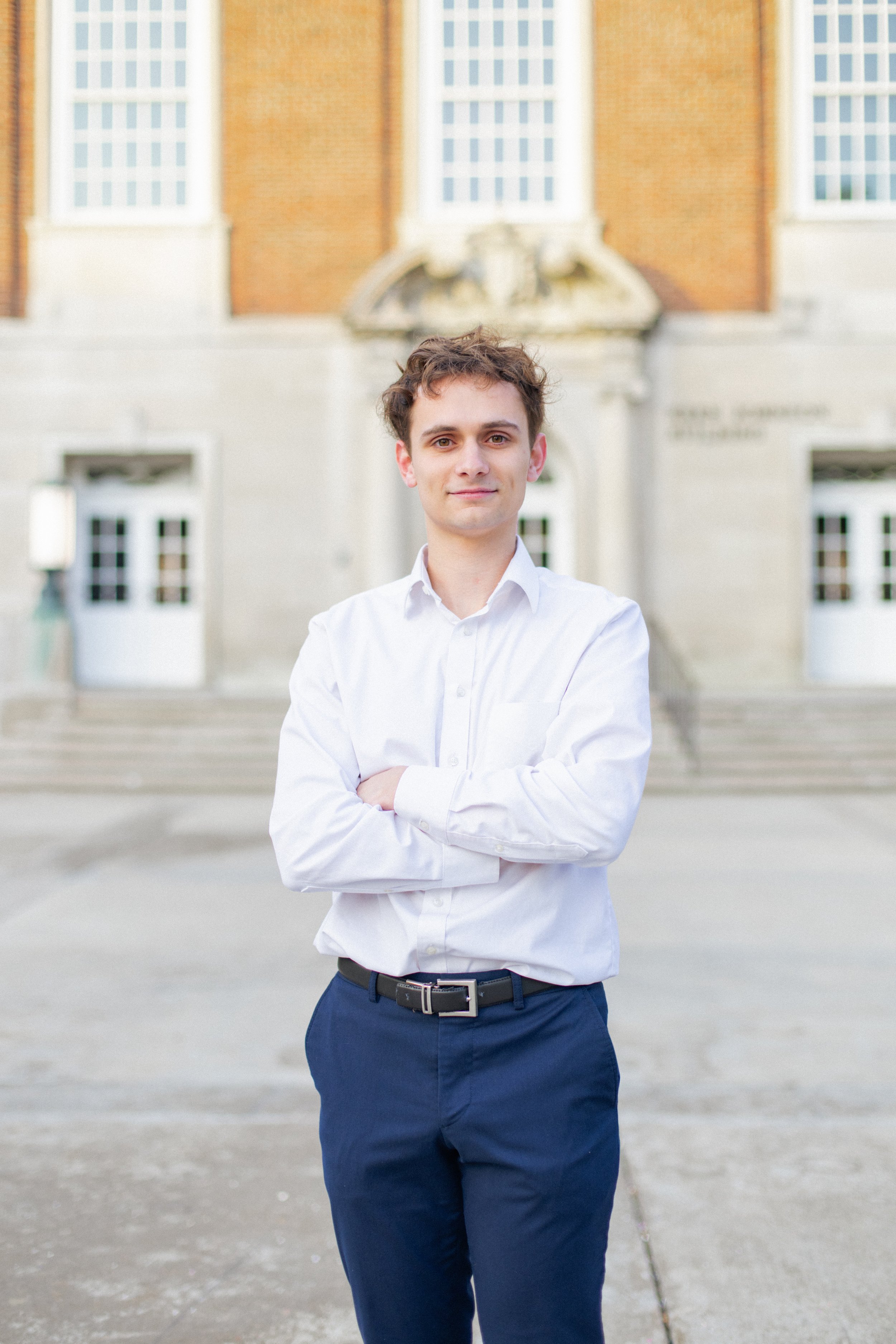 A young man in a white shirt and navy blue pants standing outside in front of a brick building with steps.