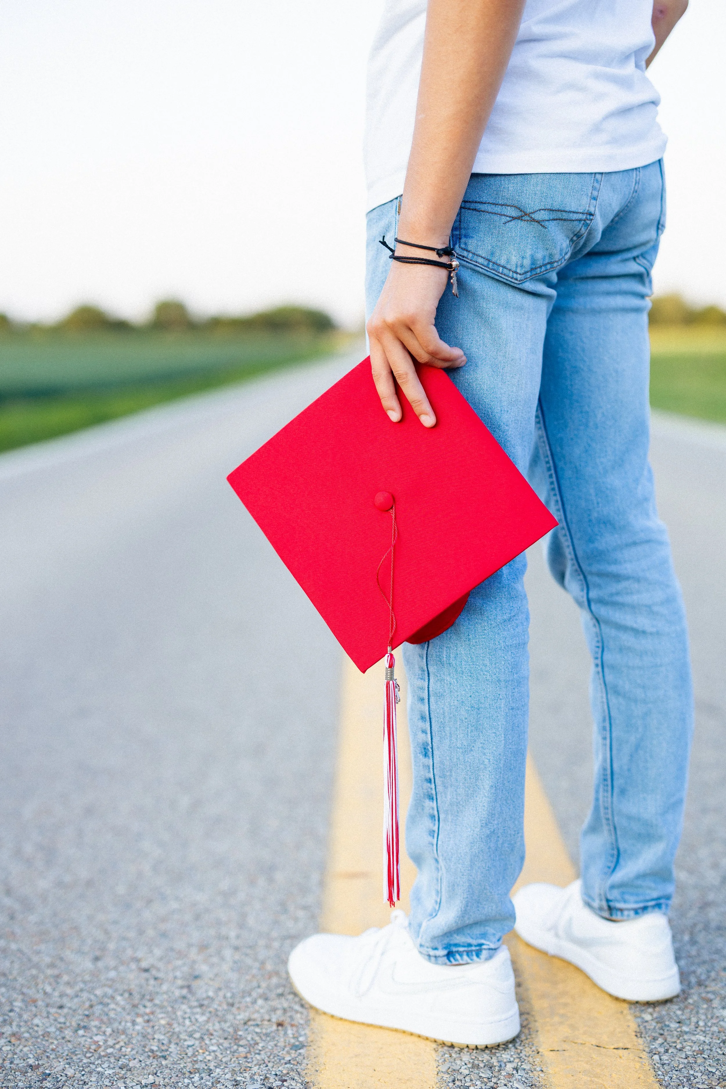 Person in jeans and sneakers holding a red graduation cap on an empty road.