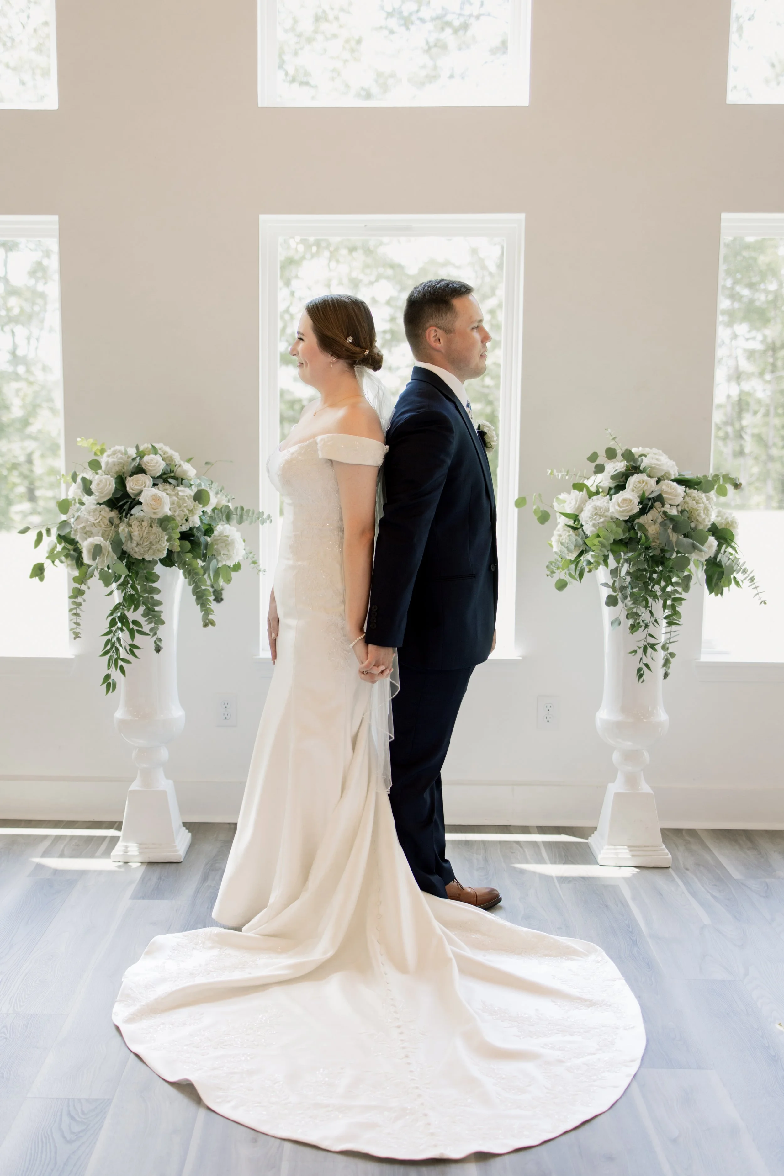 Bride and groom standing back-to-back in a wedding ceremony, holding hands, with floral arrangements on either side, in a bright room with large windows.