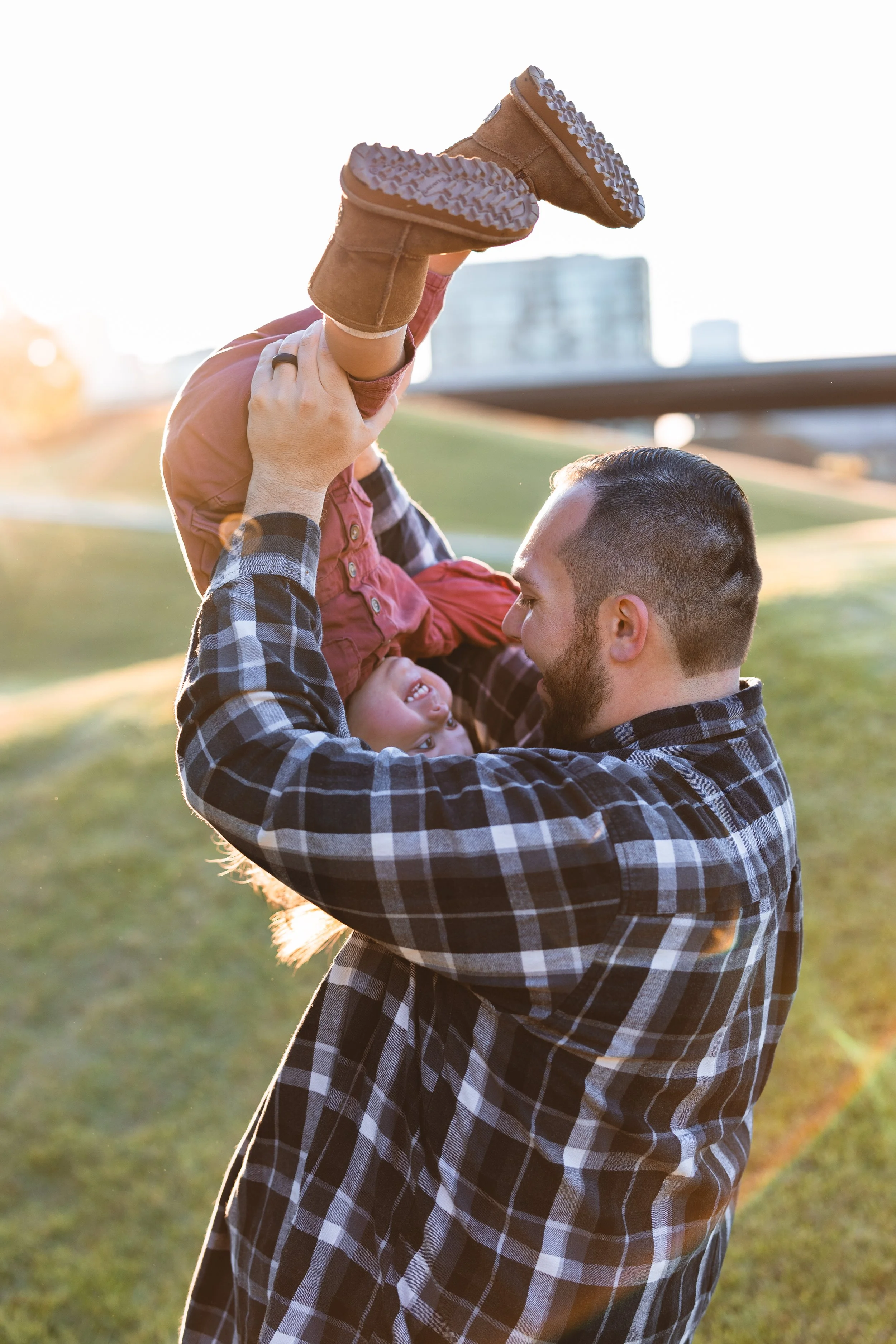 A man playing with a young girl in a park during sunset, lifting her upside down and both smiling.