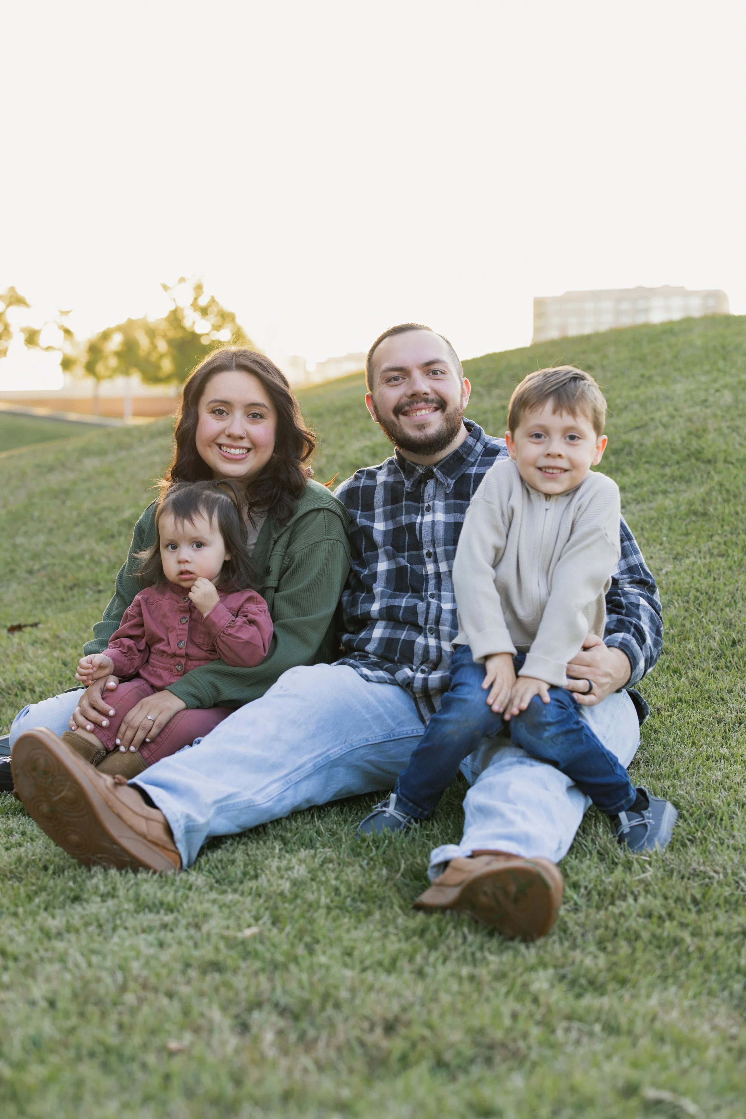 Family of four sitting on grass in a park, smiling at the camera during sunset.