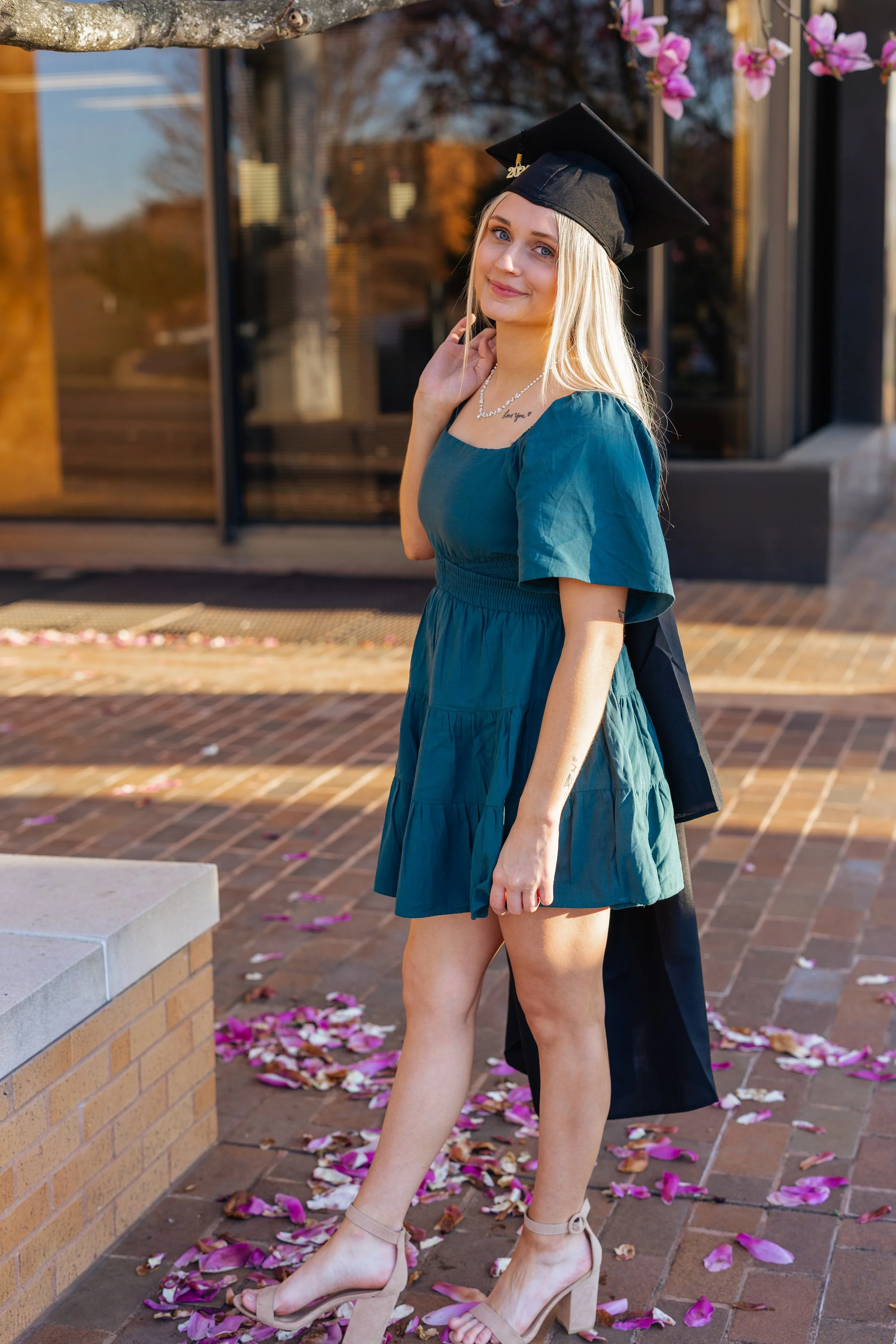 A young woman in a teal dress and tan heels is wearing a black graduation cap. She is standing outside on a brick sidewalk with pink flower petals scattered around, near a tree with pink blossoms. She has long blonde hair and is smiling softly at the
