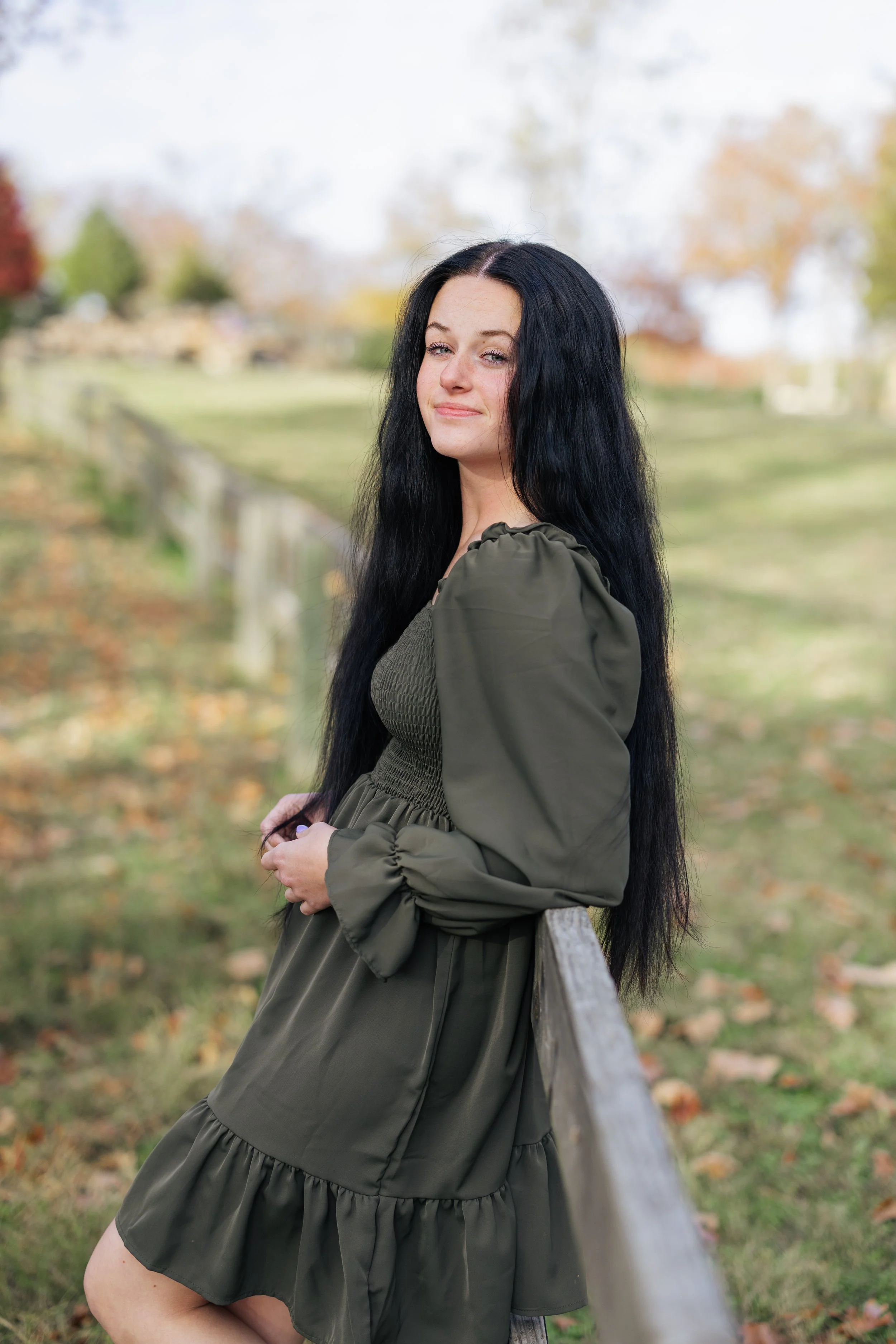 A young woman with long black hair wearing an olive green dress with puffed sleeves, standing outdoors in a park during fall, leaning against a wooden fence.