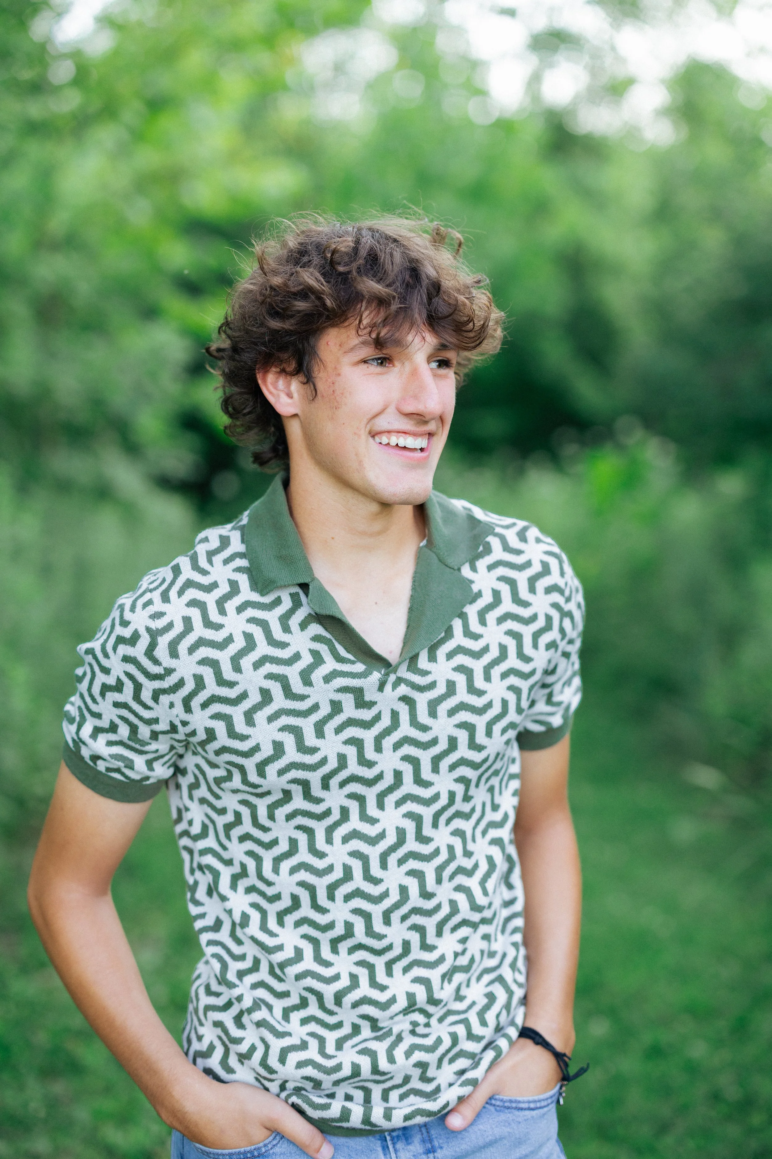 A young man with curly brown hair and a bright smile stands outdoors in front of green trees, wearing a patterned green and white polo shirt and blue jeans.