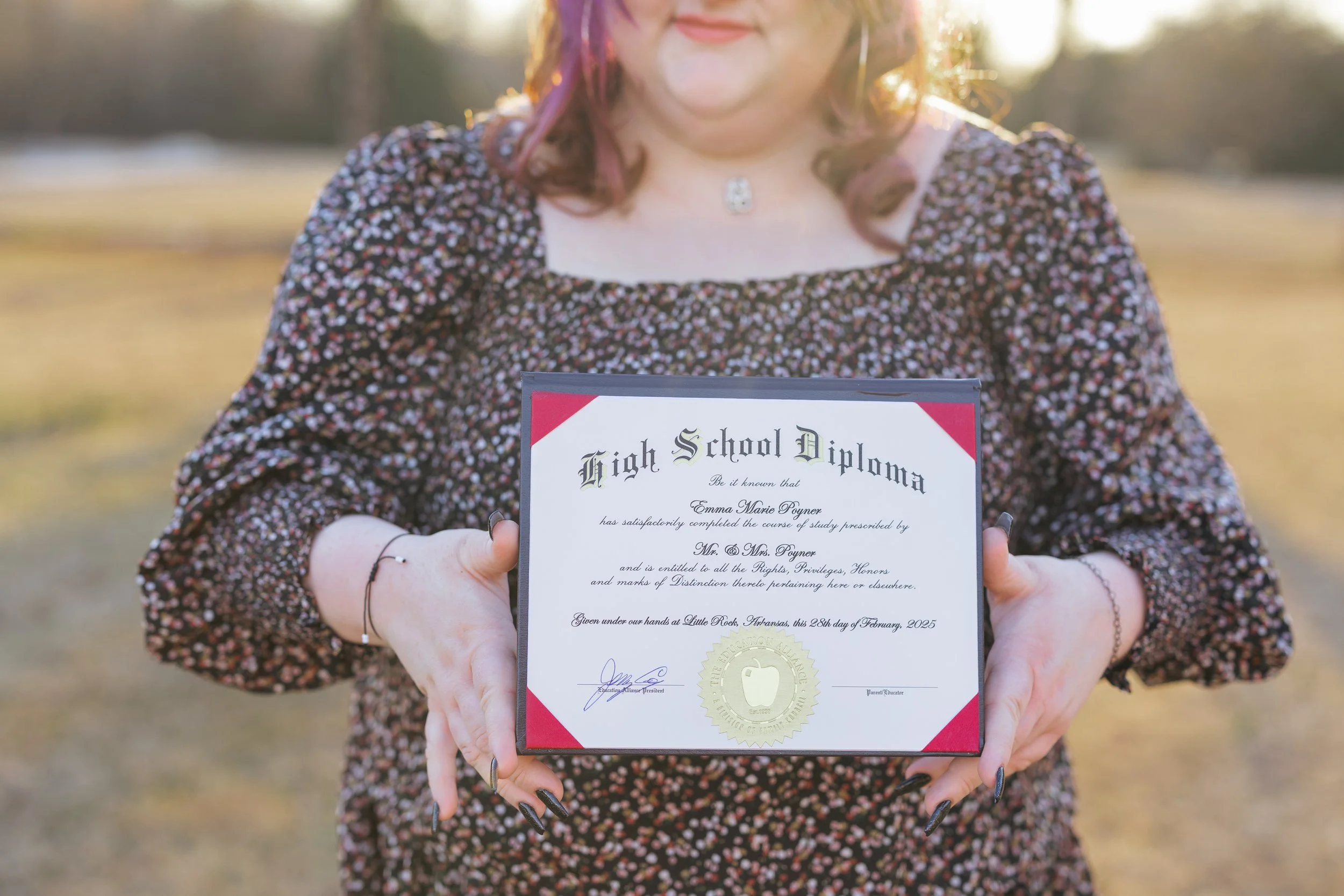 Woman holding a high school diploma certificate outdoors in a field during sunset.