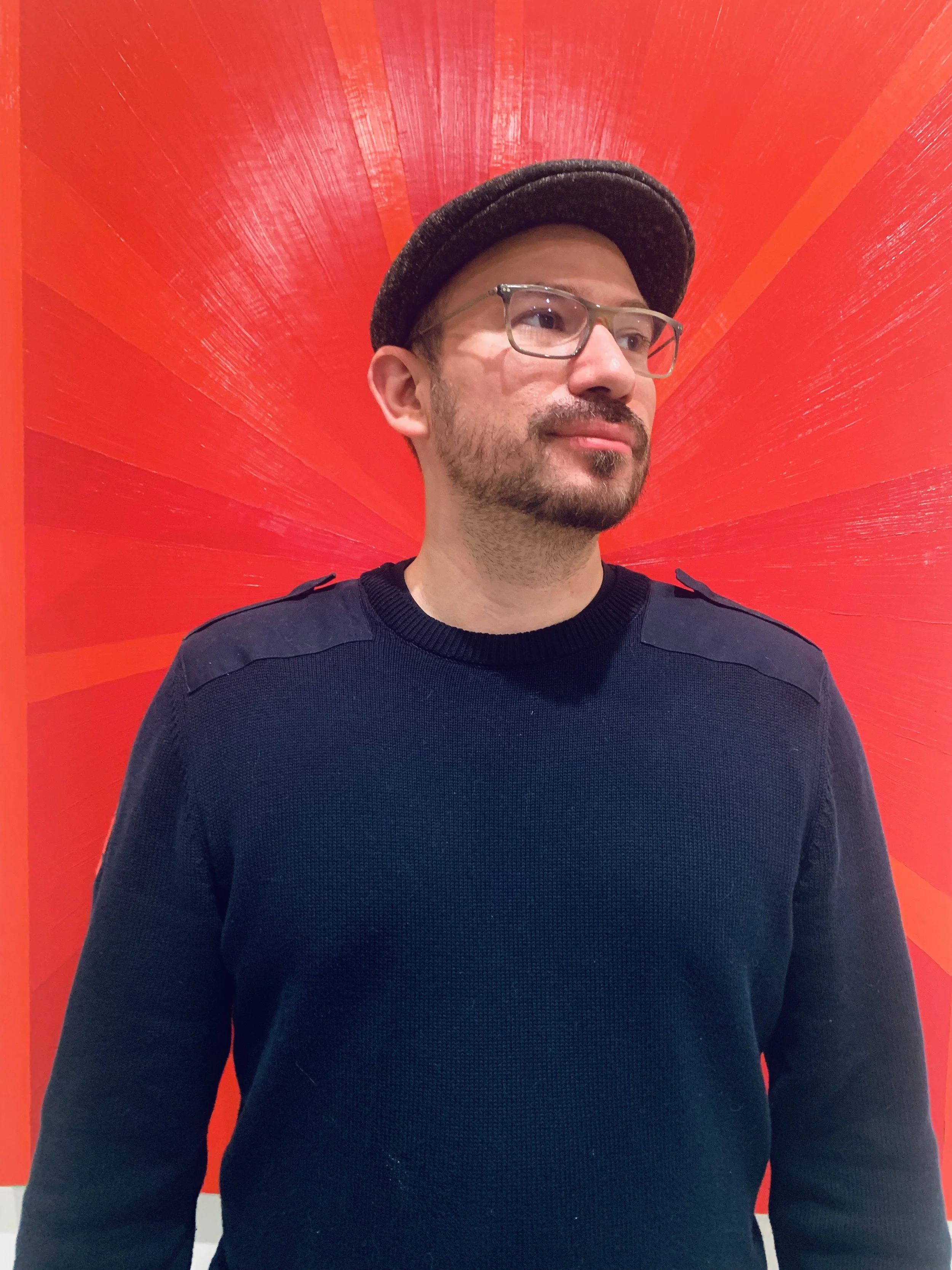 Man wearing glasses, Victor Hugo Orozco, and a cap standing in front of a bright red background with a starburst pattern.