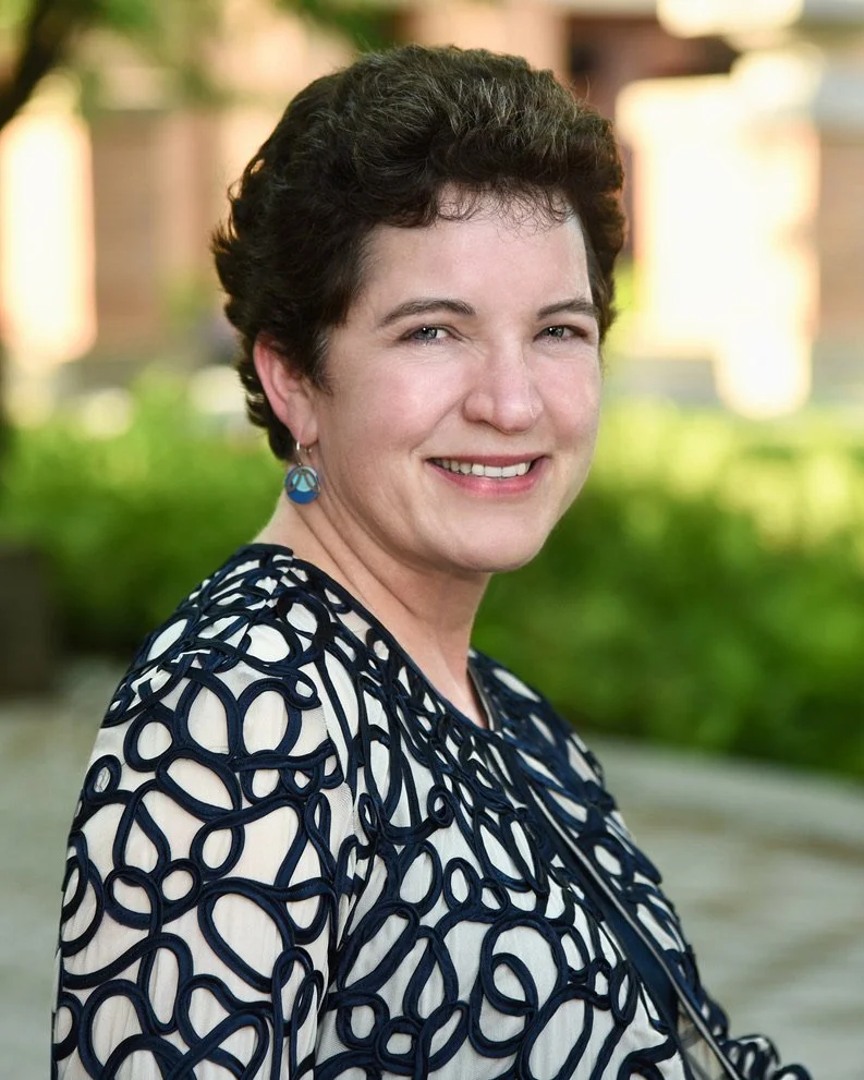 Smiling woman Kim Marie McKernan, with short curly hair, wearing a patterned top and earrings, standing outdoors with greenery in the background.