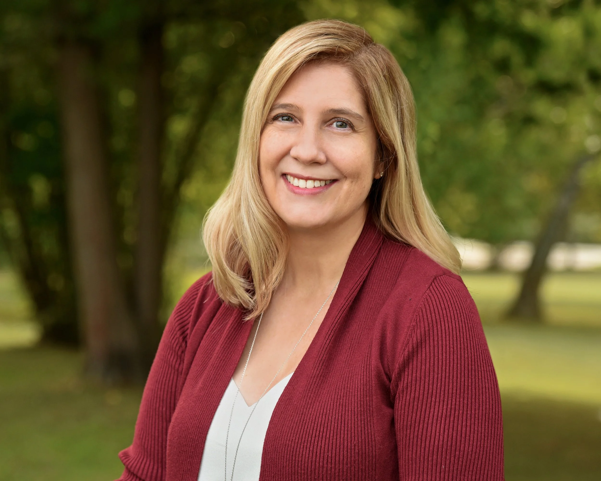 Smiling woman, Cassie Conroy, in a red sweater with blonde hair, standing outdoors with trees in the background.