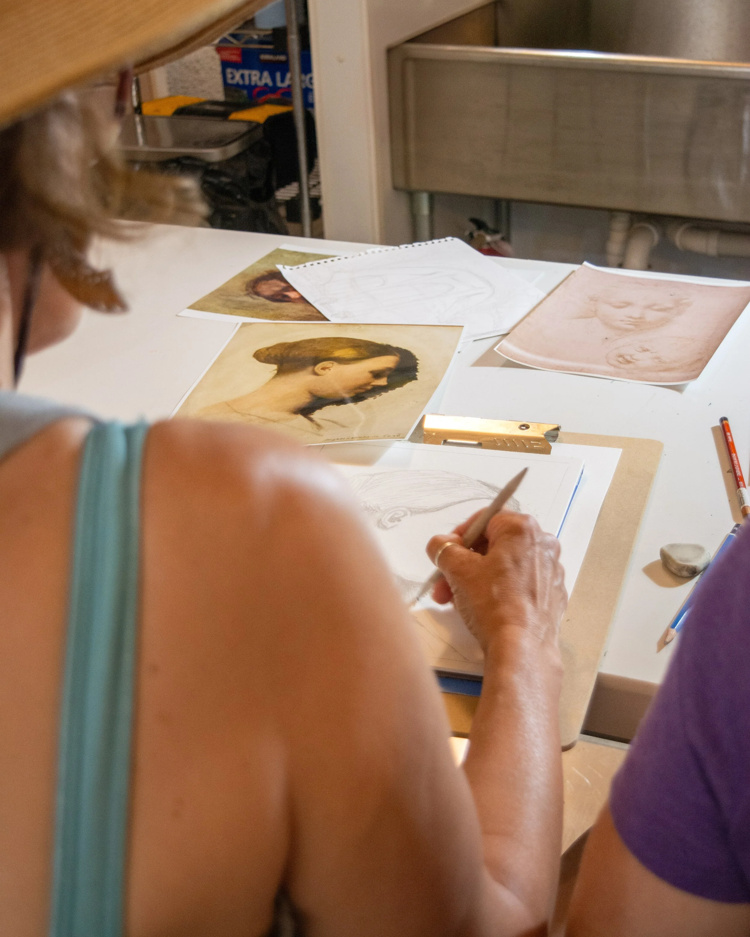 An artist sketches a portrait at a desk, with reference images of a woman and a drawing of a face on the table, and various art supplies nearby.