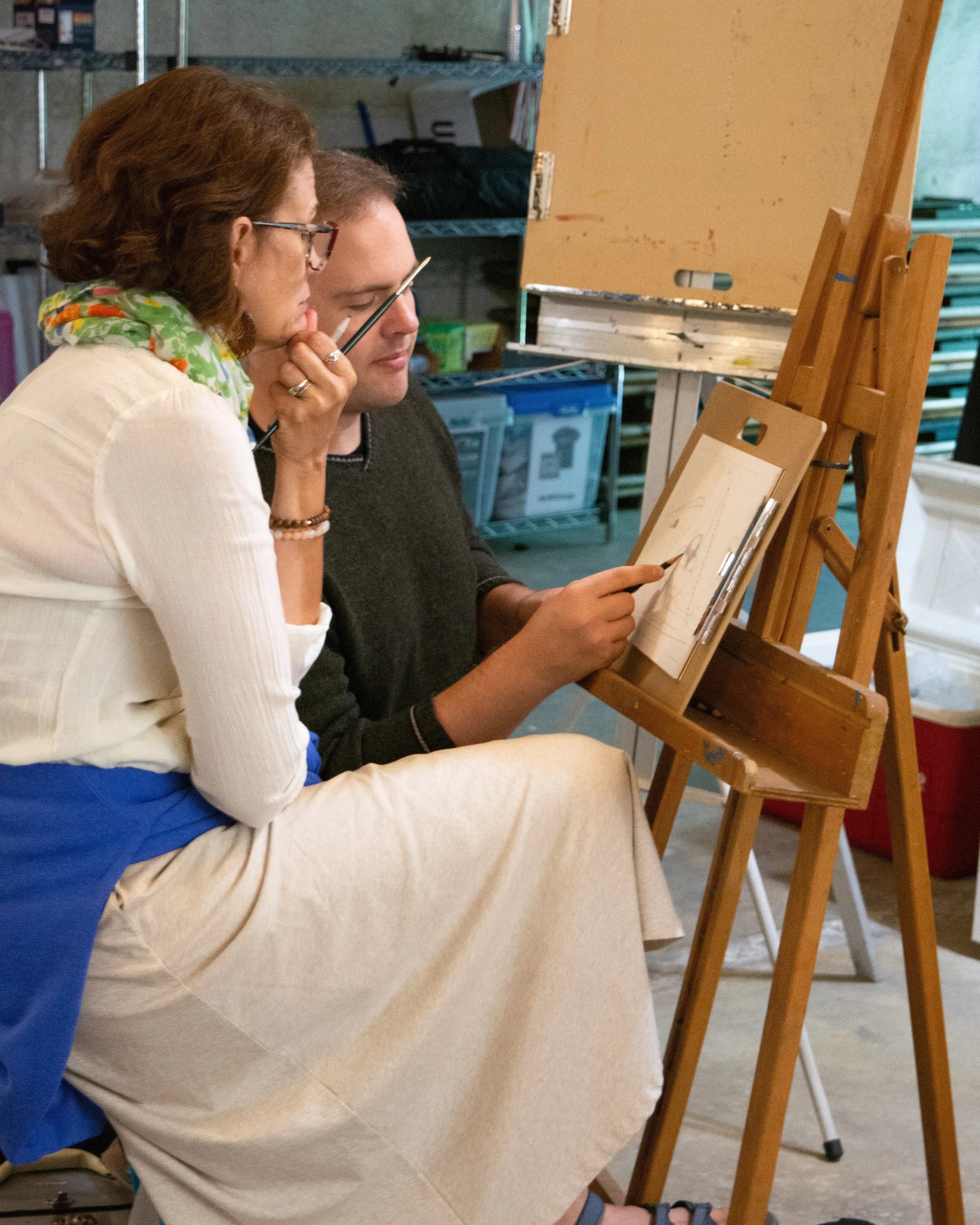 A woman and a man sitting closely together, observing a drawing on an easel, with the woman holding a paintbrush.