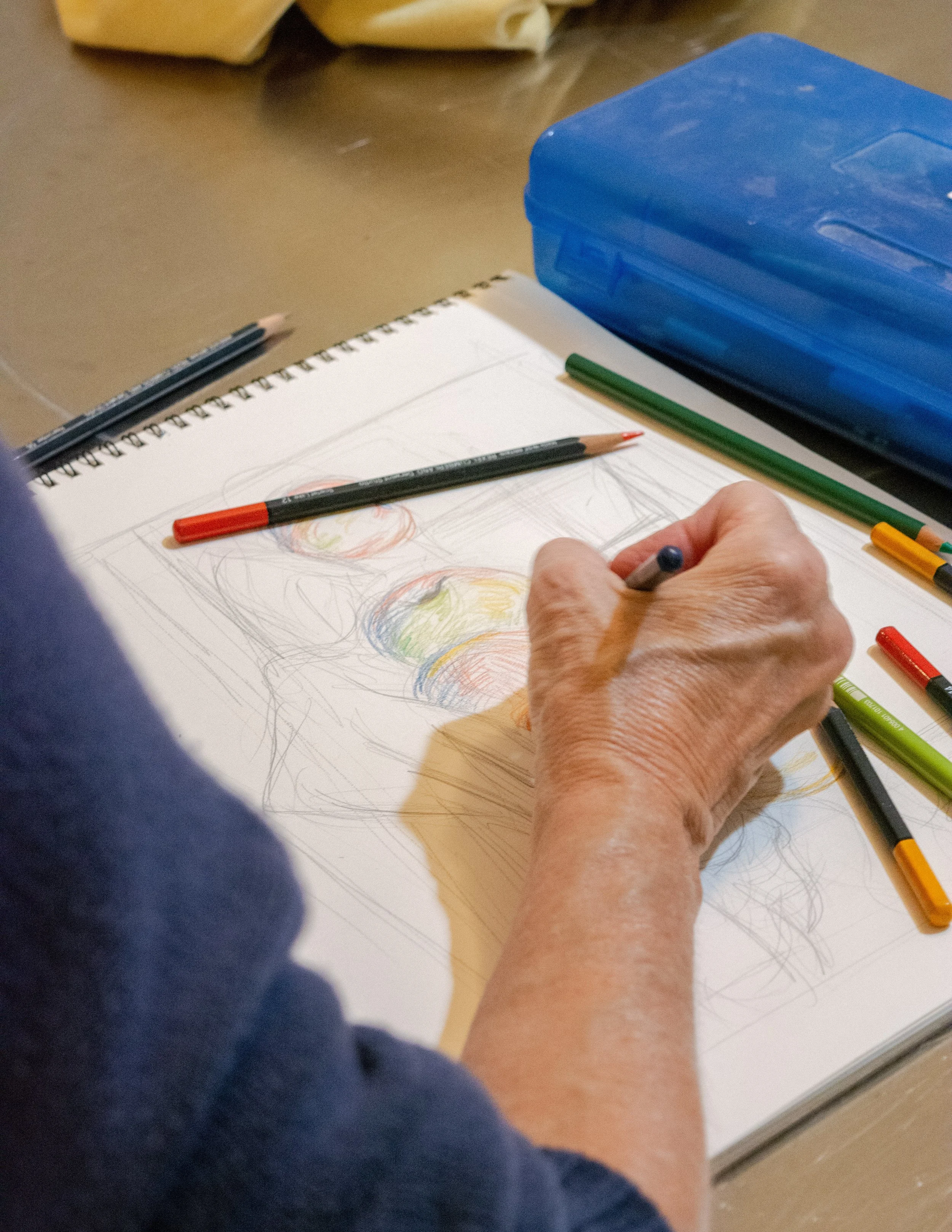 An elderly person's hand sketching colorful fruit on a spiral notebook, with colored pens and markers nearby on a desk.