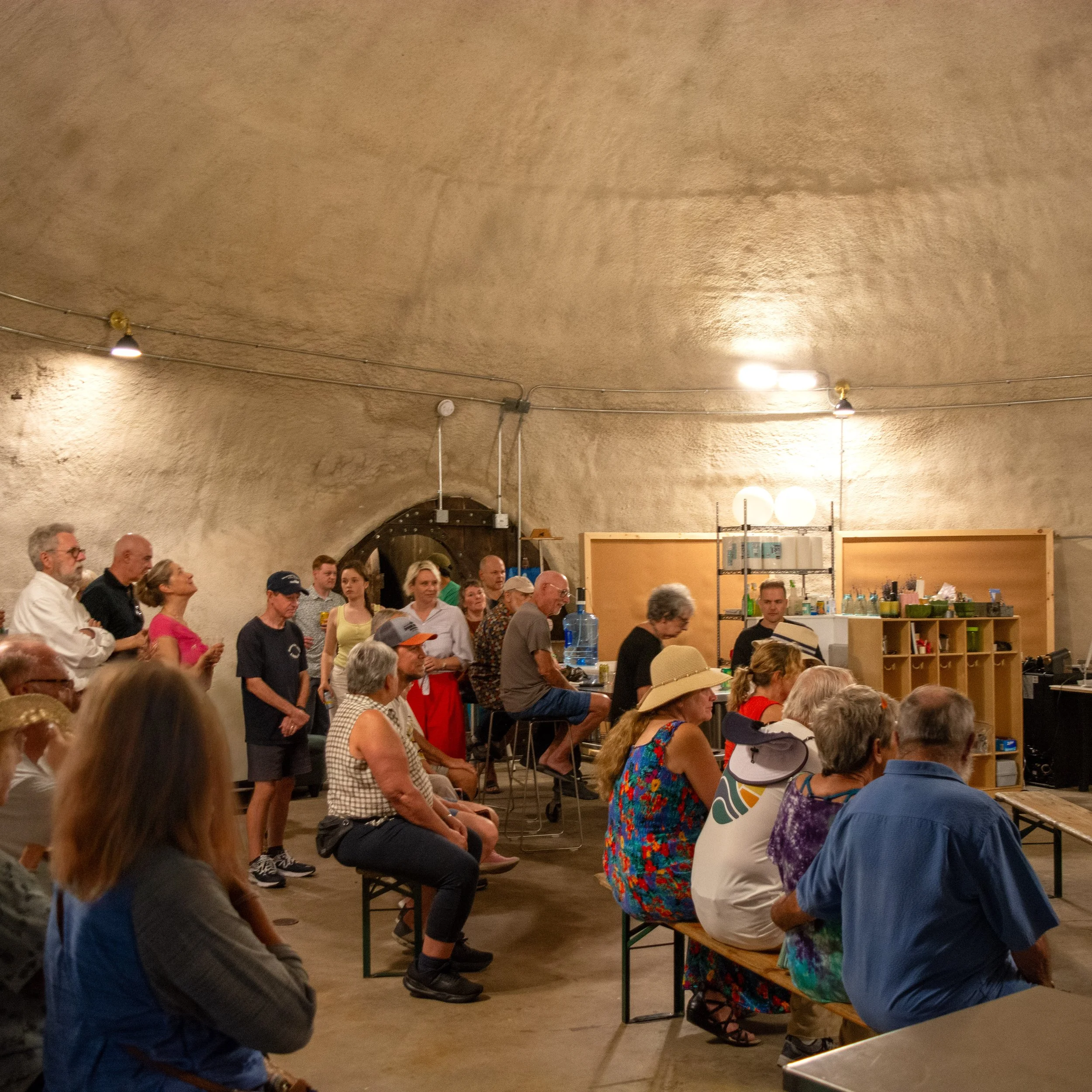 Group of people gathered in an underground room with high, curved ceiling, sitting on benches and standing, watching a presentation or performance at the front.