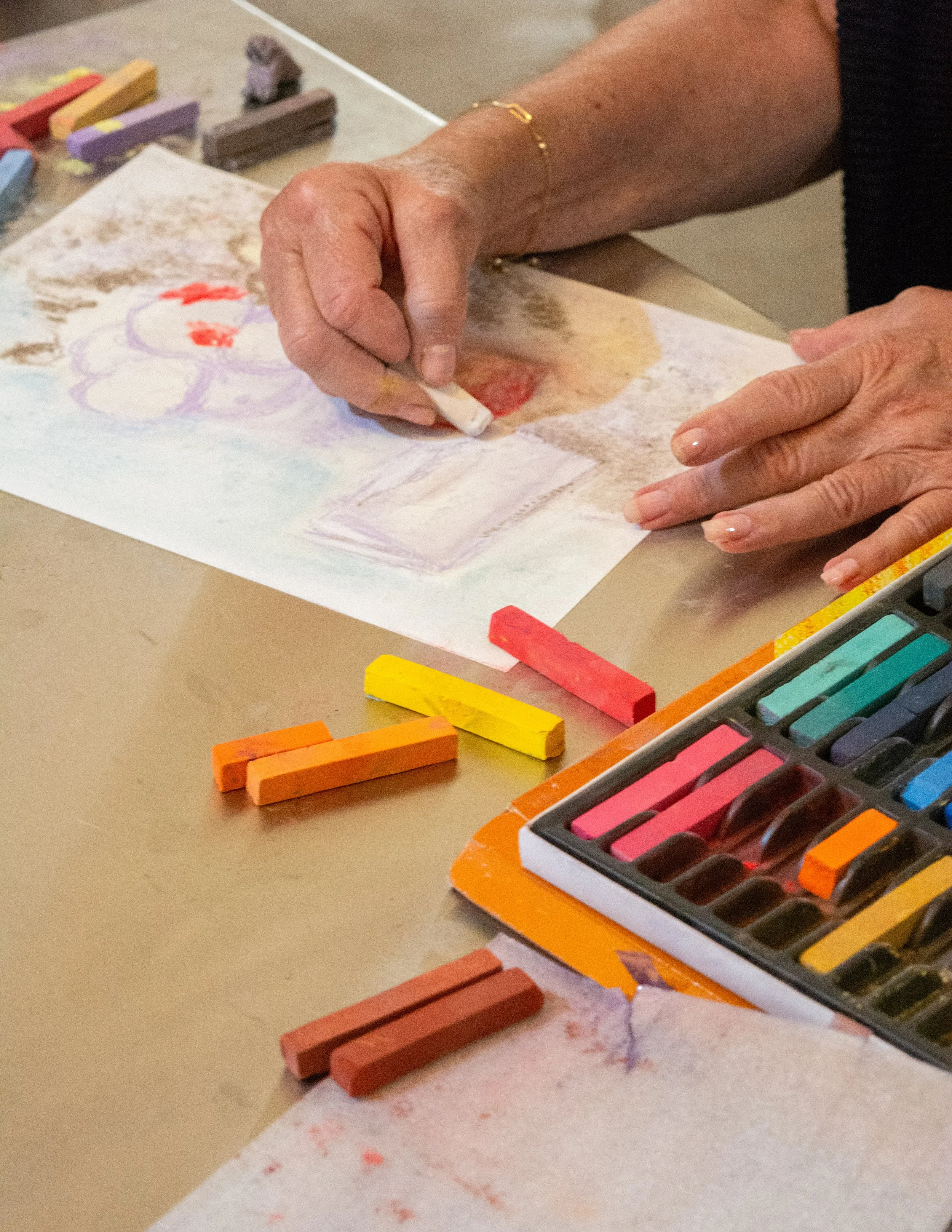 Close-up of hands using a pastel crayon to create artwork on white paper. Art supplies like colored pastels and a set of soft pastels are visible on the table at the Dome House