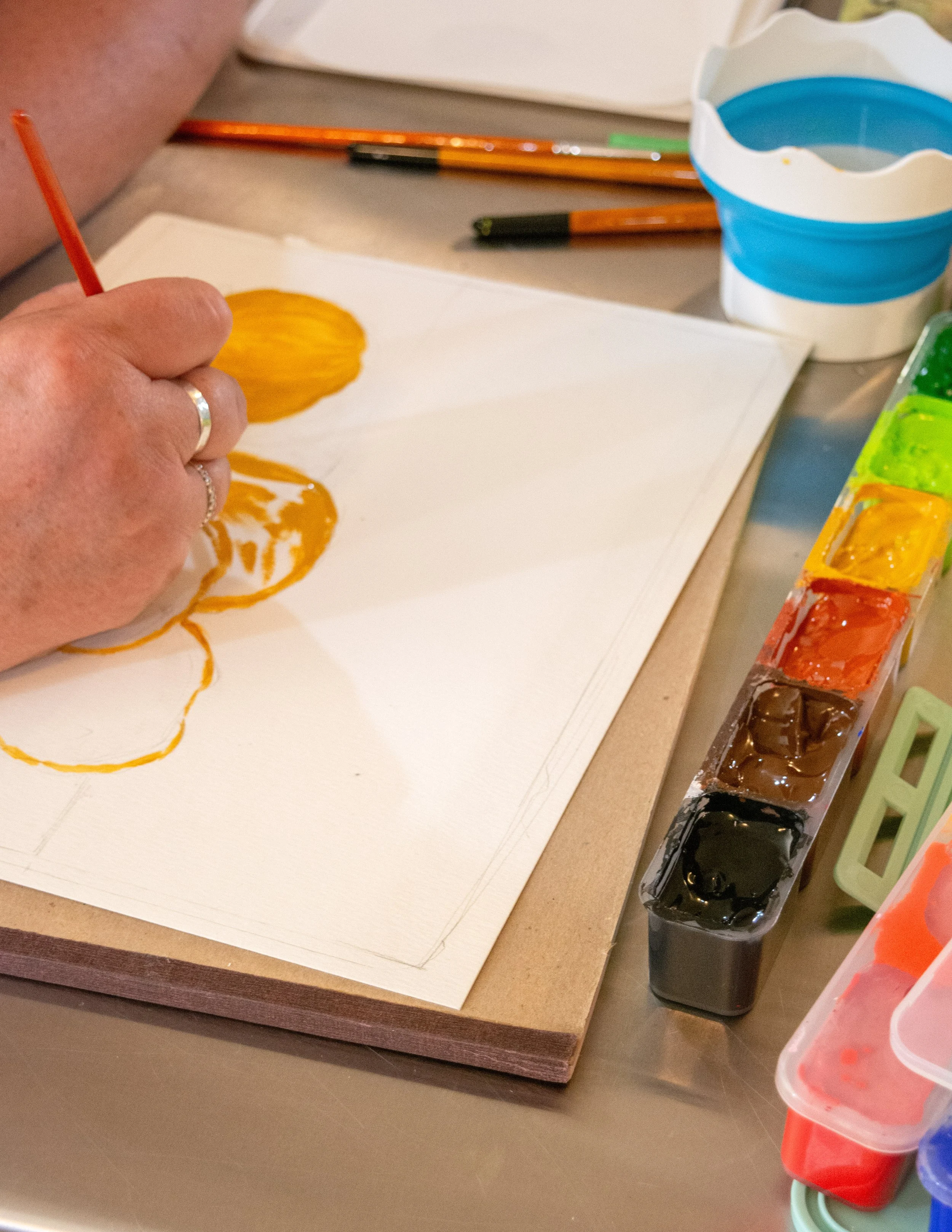 A person painting with yellow and orange watercolors on paper, with various paint supplies and brushes on the table.