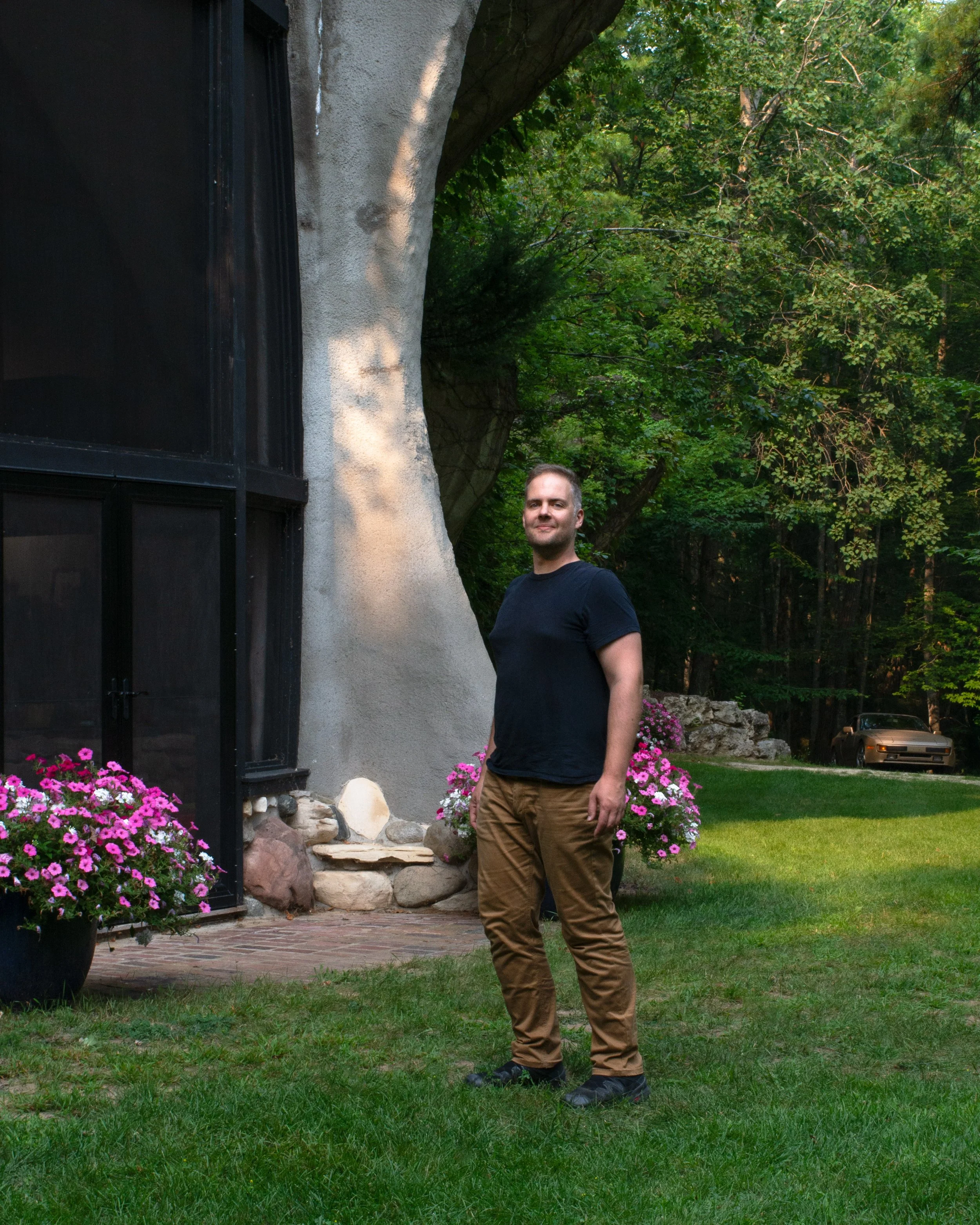 A man standing on a lawn next to a building with a concrete wall and large window, surrounded by pink flowers and trees in the background.