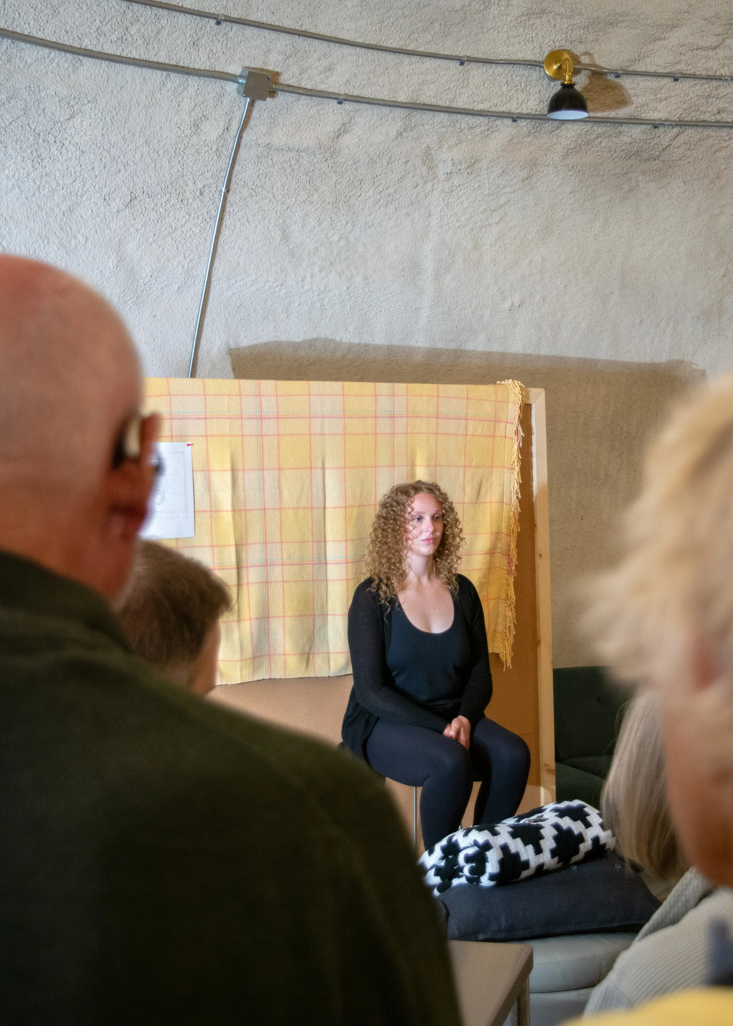 A young woman with curly hair sitting on a stool in front of an audience, appearing to perform or speak, with a beige and red checkered backdrop behind her.