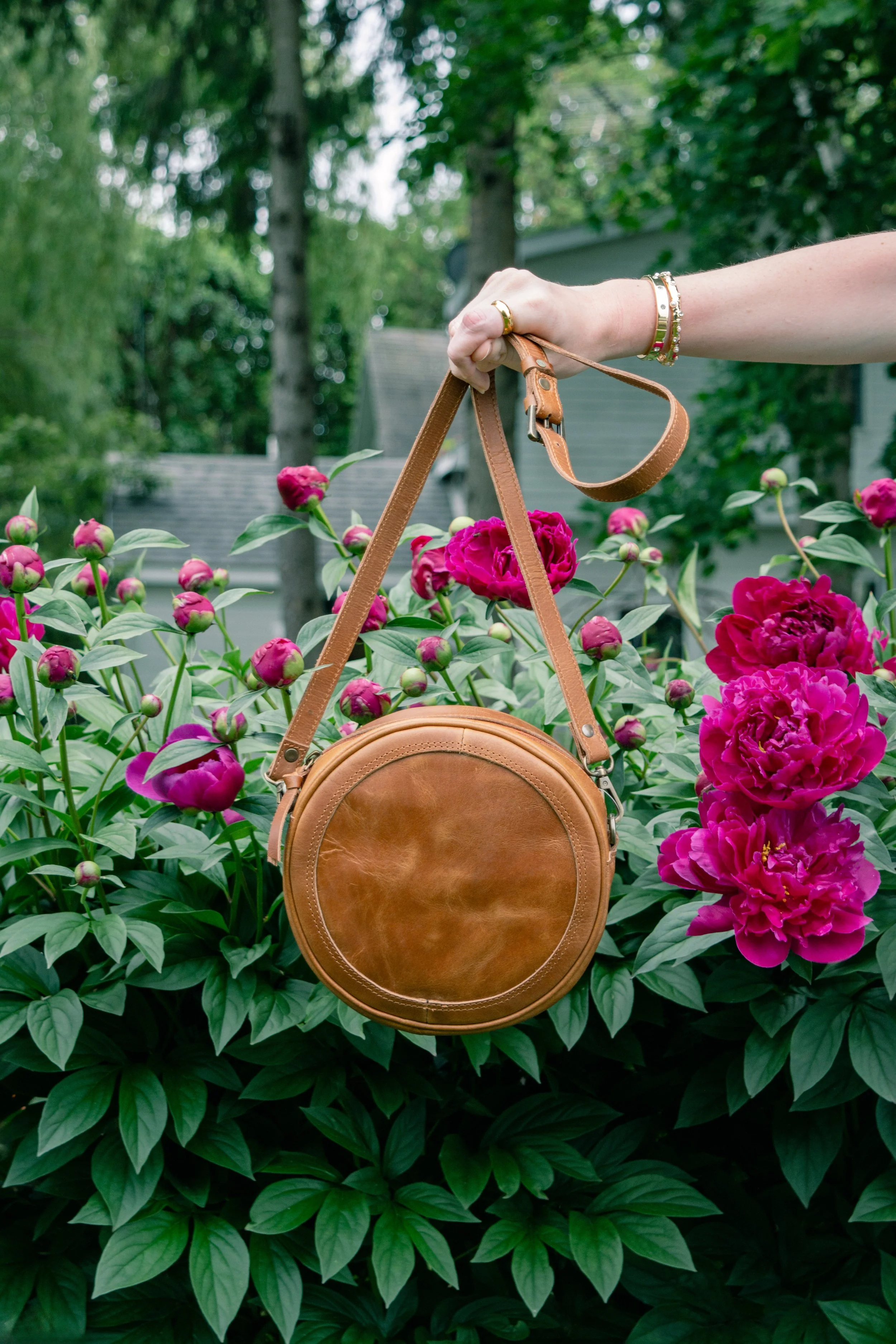 A person holding a round tan leather purse hanging from their hand, with vibrant pink peonies in full bloom in the background, surrounded by green foliage and trees.