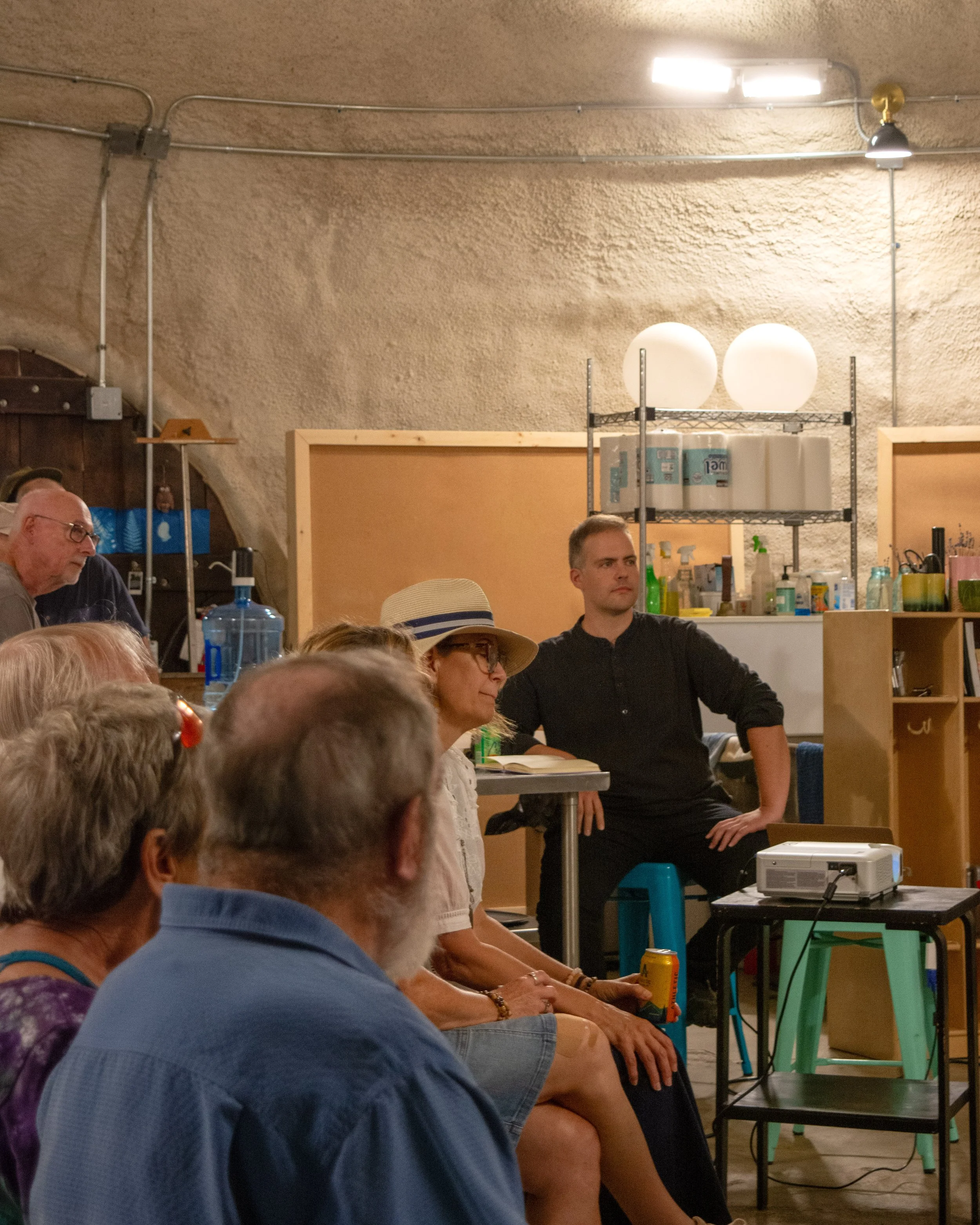 People attending a presentation or lecture in a room with textured beige walls, some seated and listening, one woman wearing a hat, another man in a black shirt, a projector on a small table, and various shelves with items in the background.