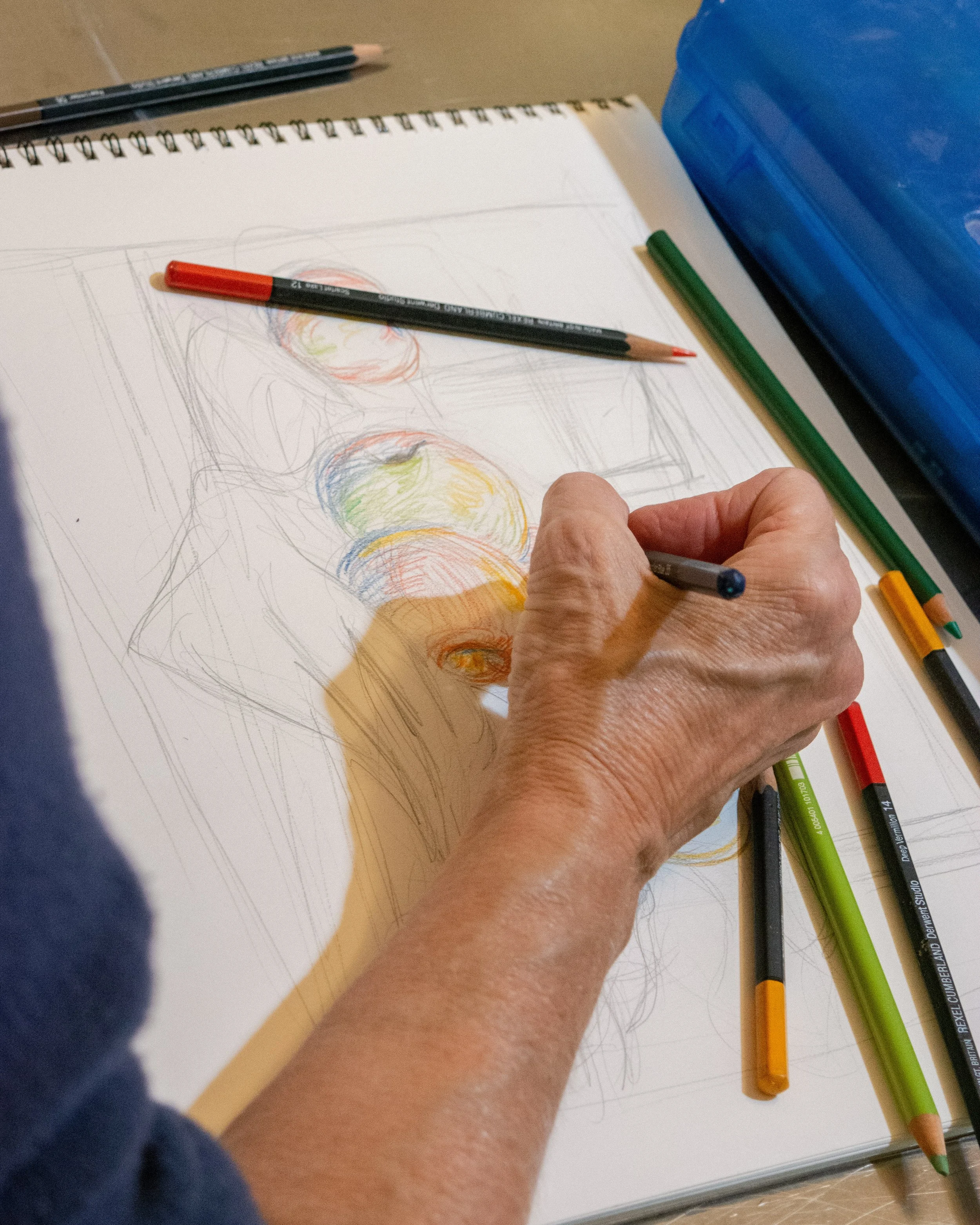 A person drawing a colorful still life on a sketchpad, surrounded by colored pencils and pens.