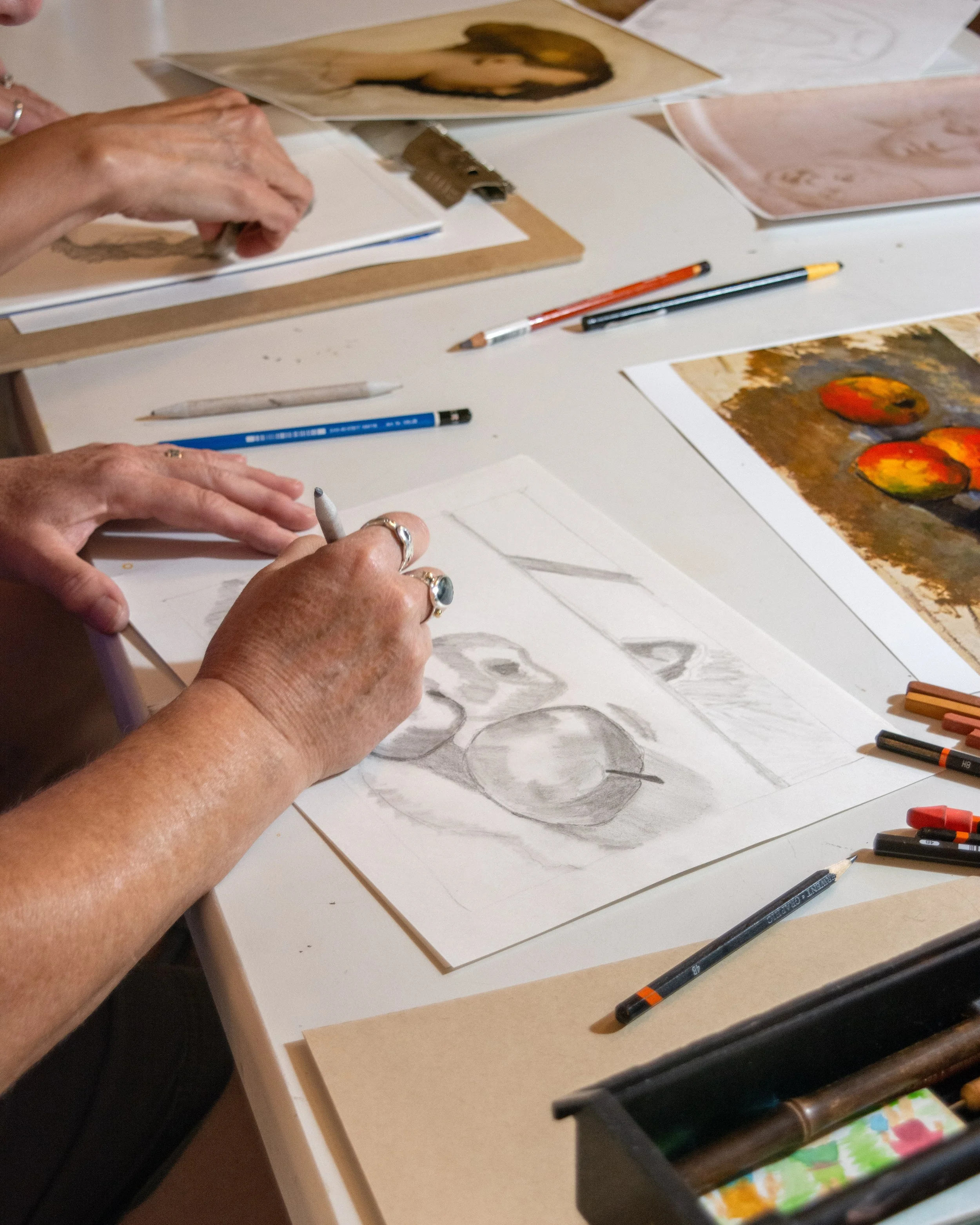 People working on art projects with drawings, paintings, and art supplies on a white table.
