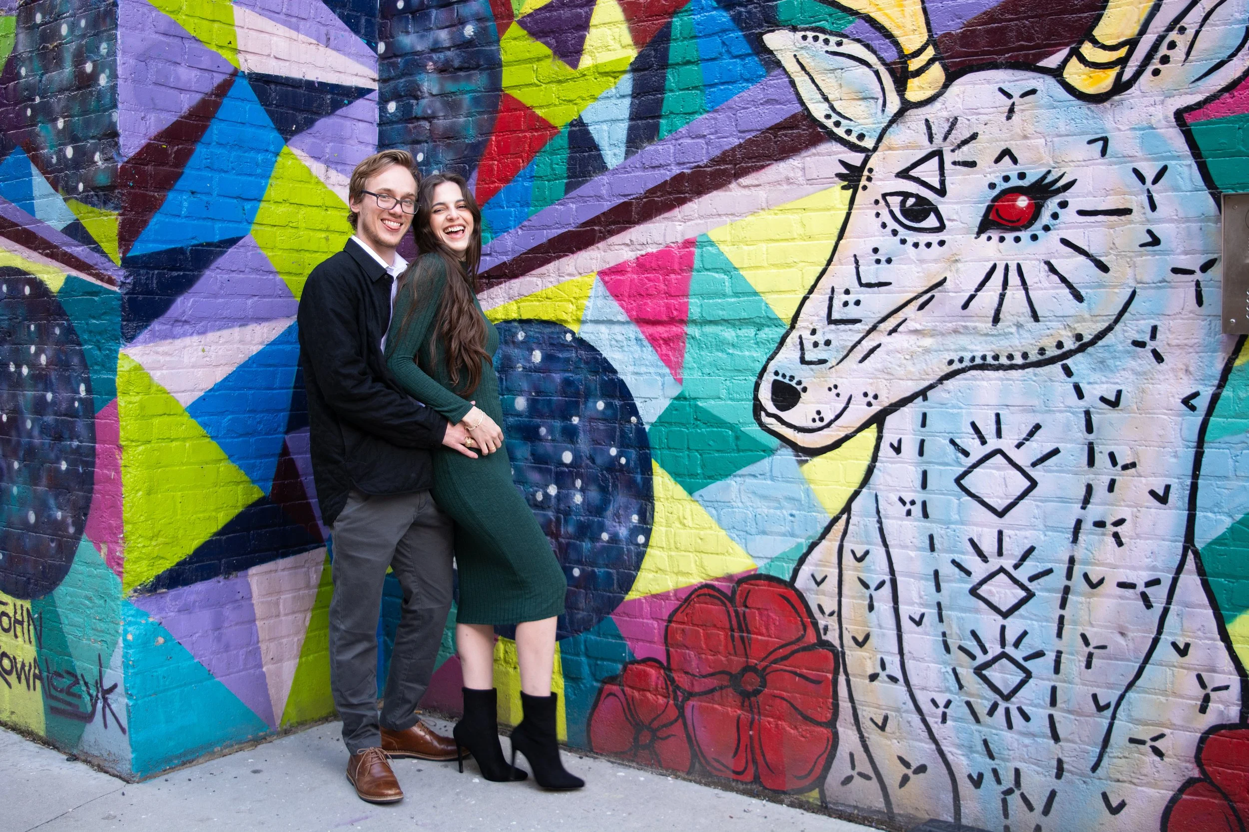 A happy couple standing in front of a colorful graffiti mural of a unicorn with red flowers on the wall.