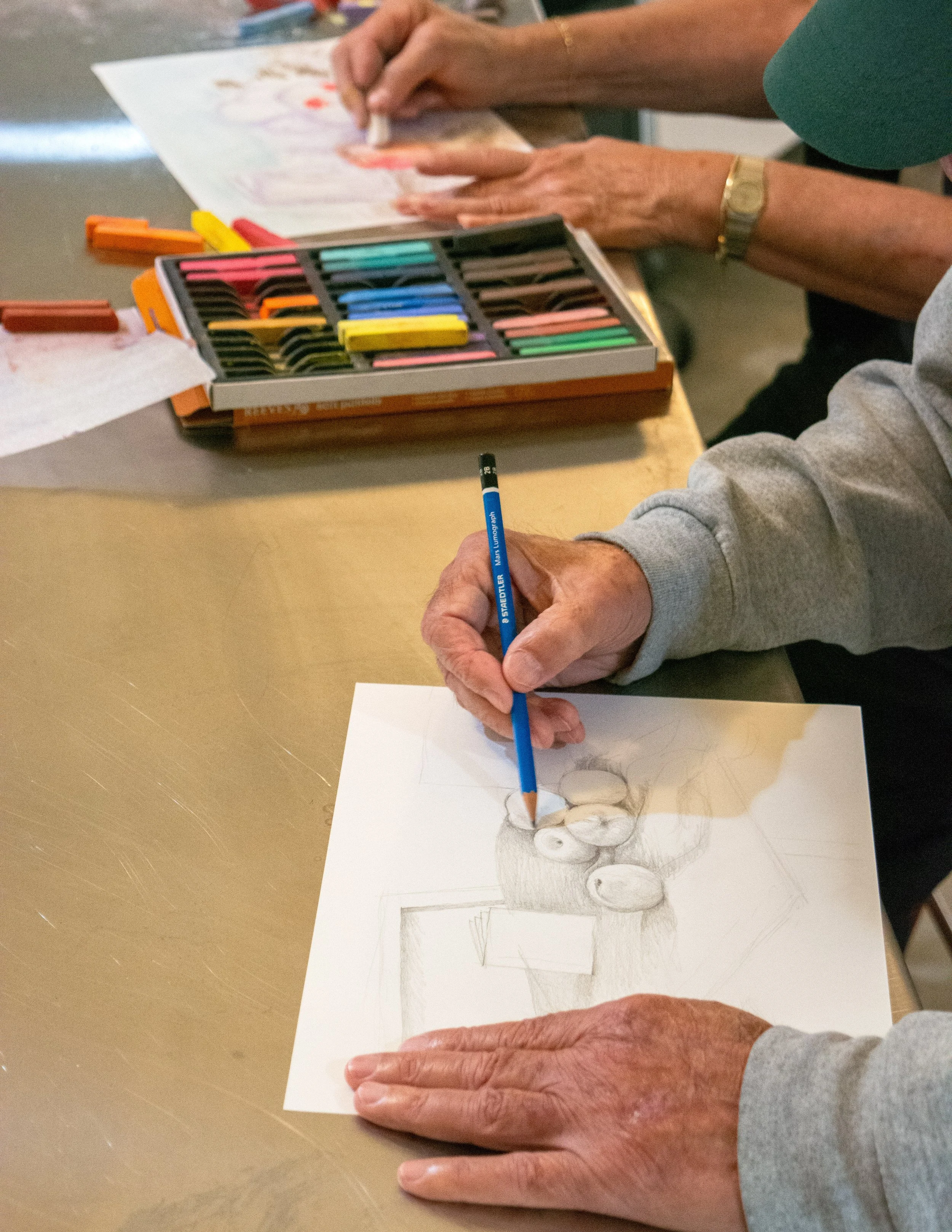 Two people drawing and coloring with pastels and pencils, with a box of colorful pastels on the table at the Dome House