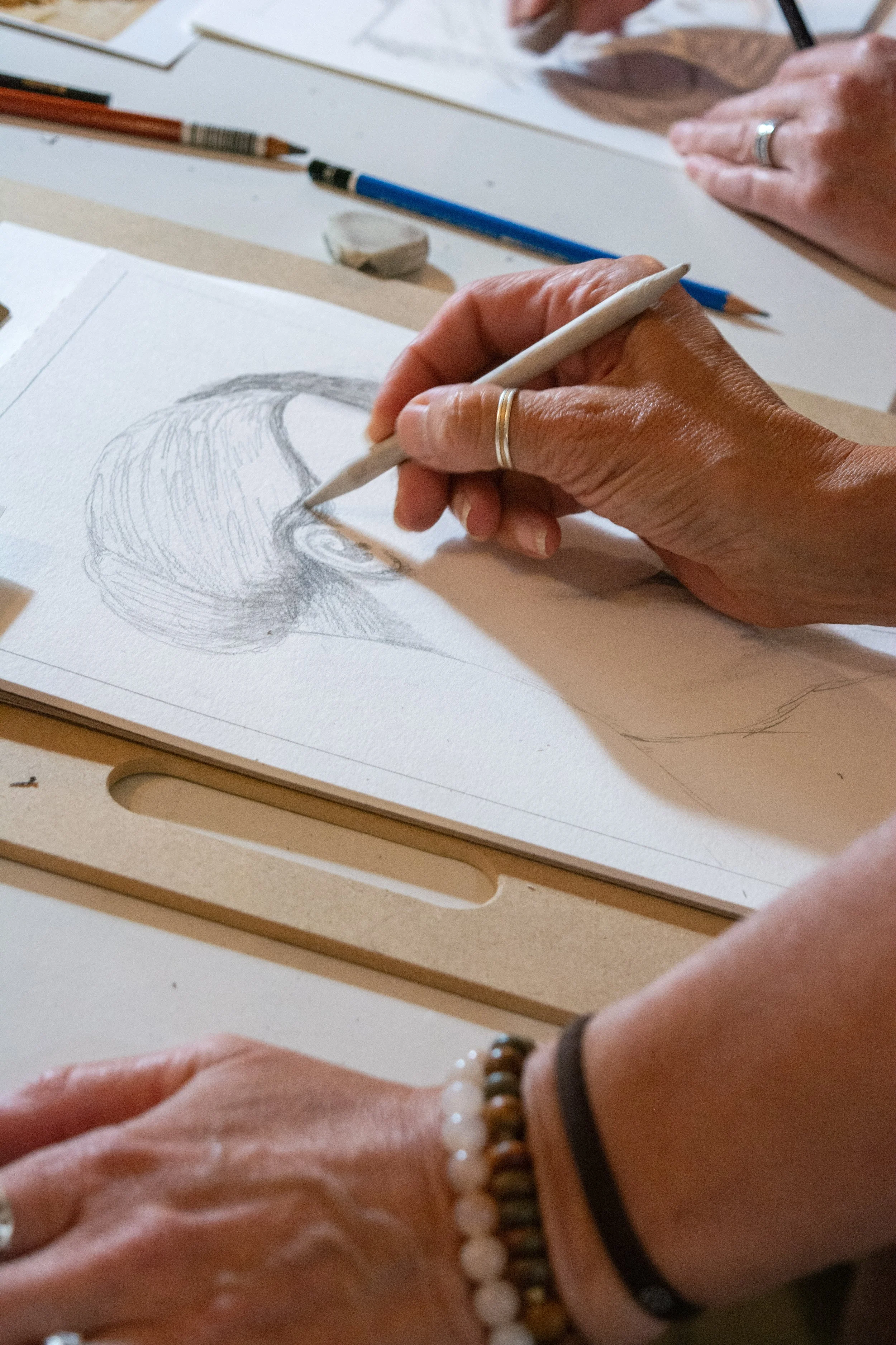 A person sketching a portrait with a pencil on a white drawing paper, others observing the artwork, with art supplies and erasers on the table.