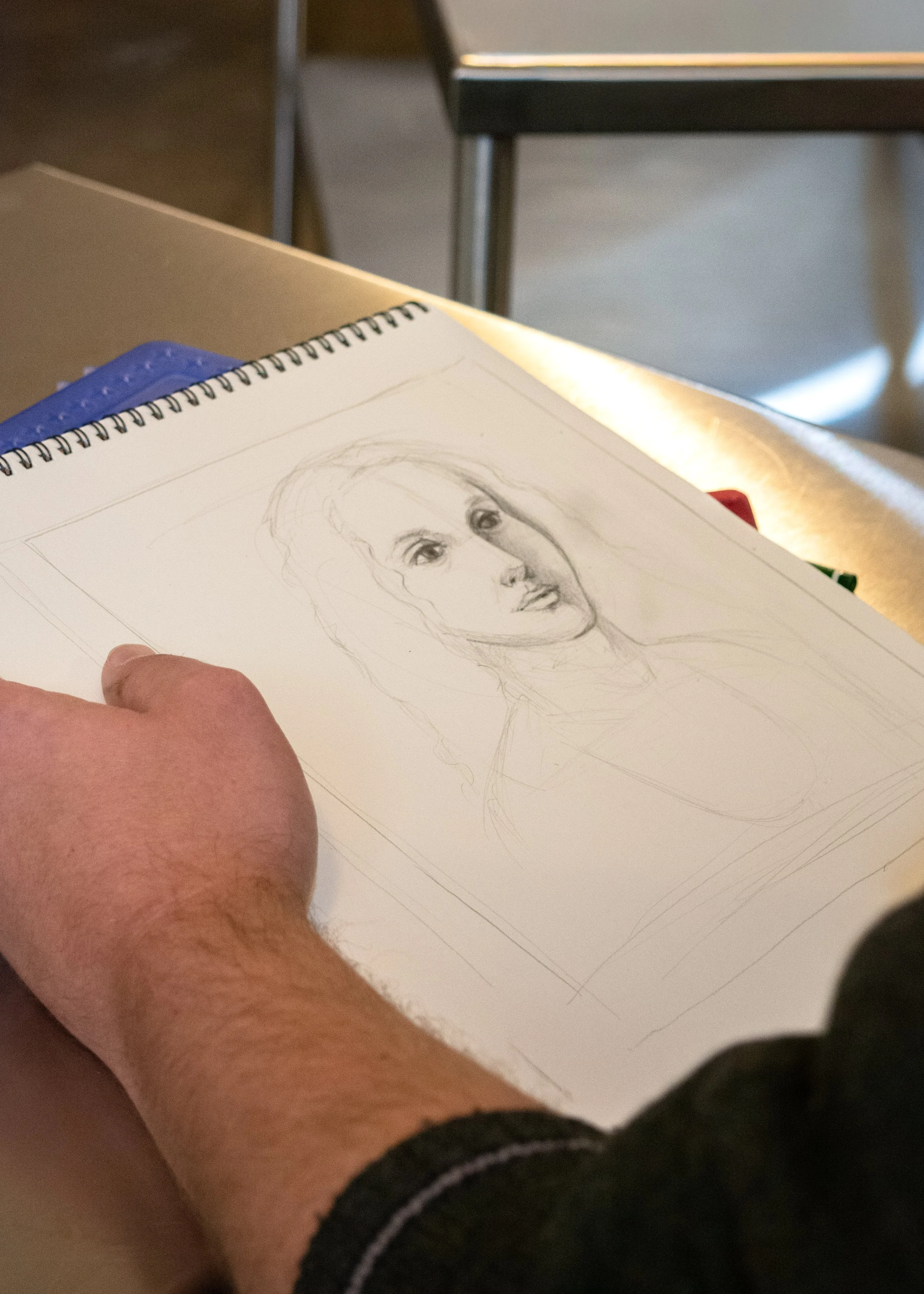 A sketch of a woman's portrait on a drawing pad, with a person's hand visible in the foreground, and a chair and part of a table in the background.