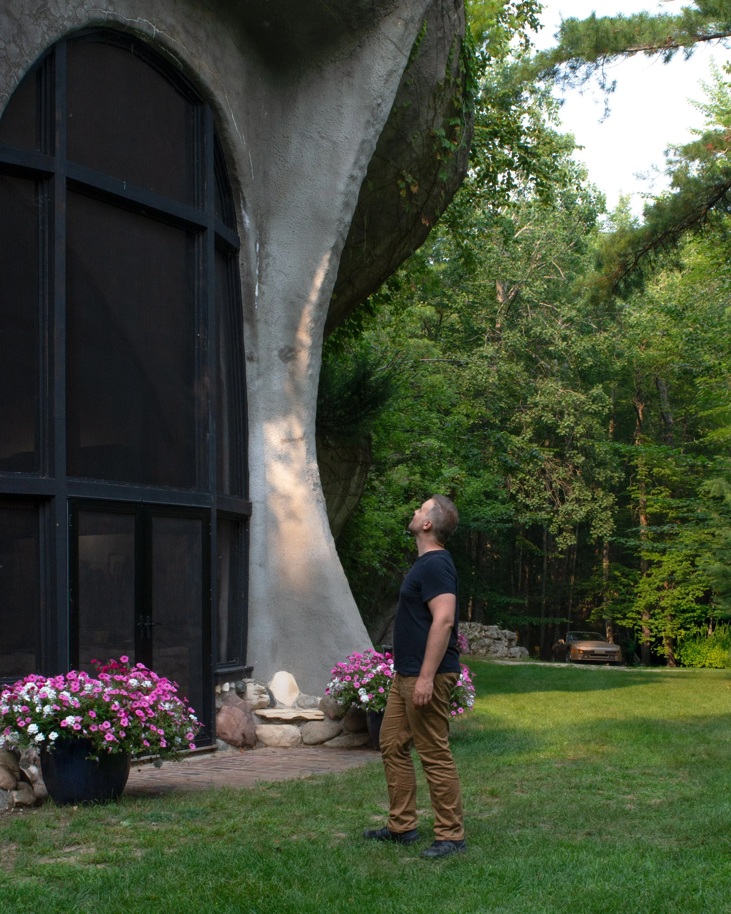 A man with short hair and a beard standing on a lawn, looking up at a large, uniquely shaped building with a rounded, overhanging roof and black framed windows, surrounded by pink and white flowers and trees.