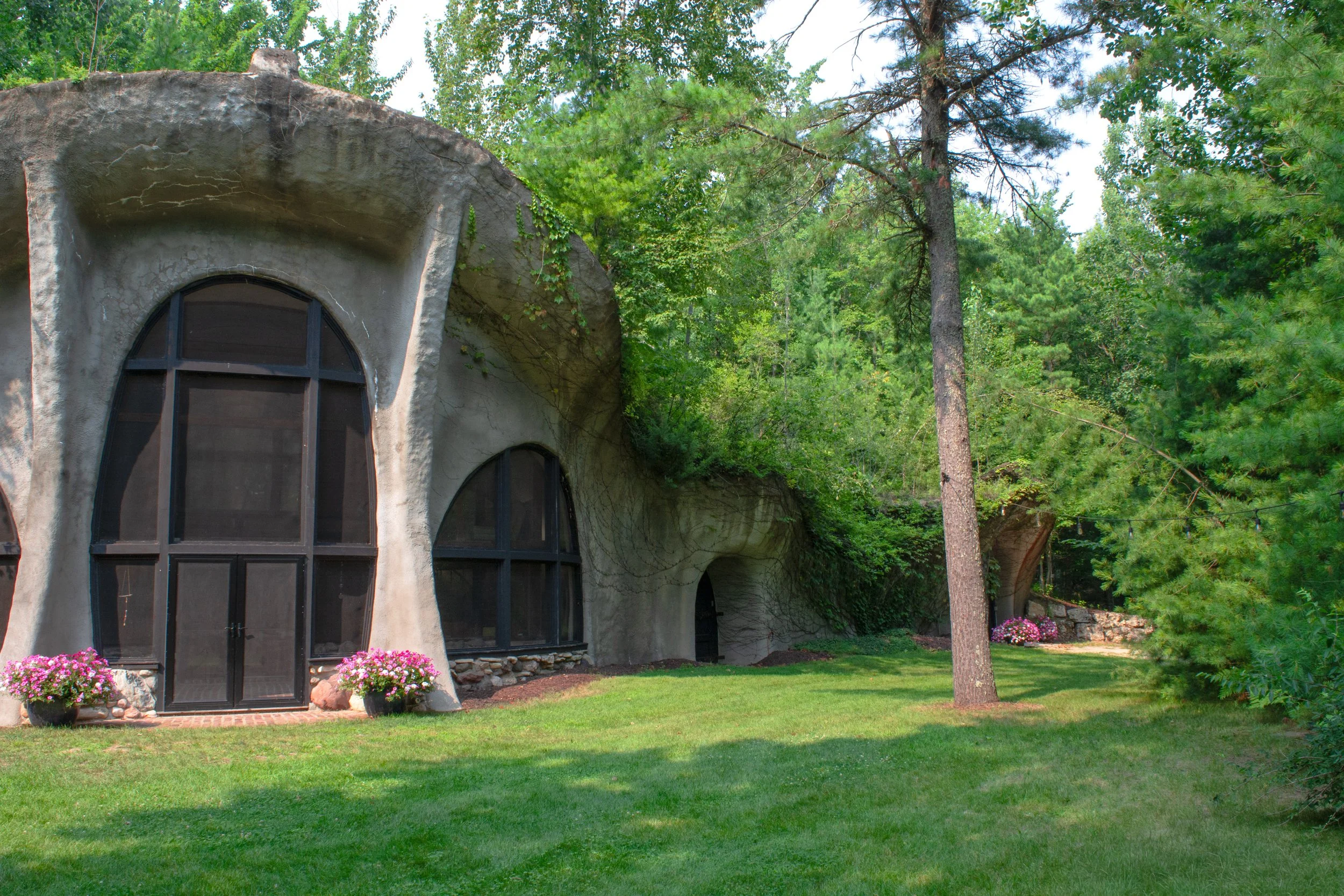 A house with rounded, irregular concrete walls and large curved windows, surrounded by a well-maintained grassy yard, trees, and flower pots with pink flowers.